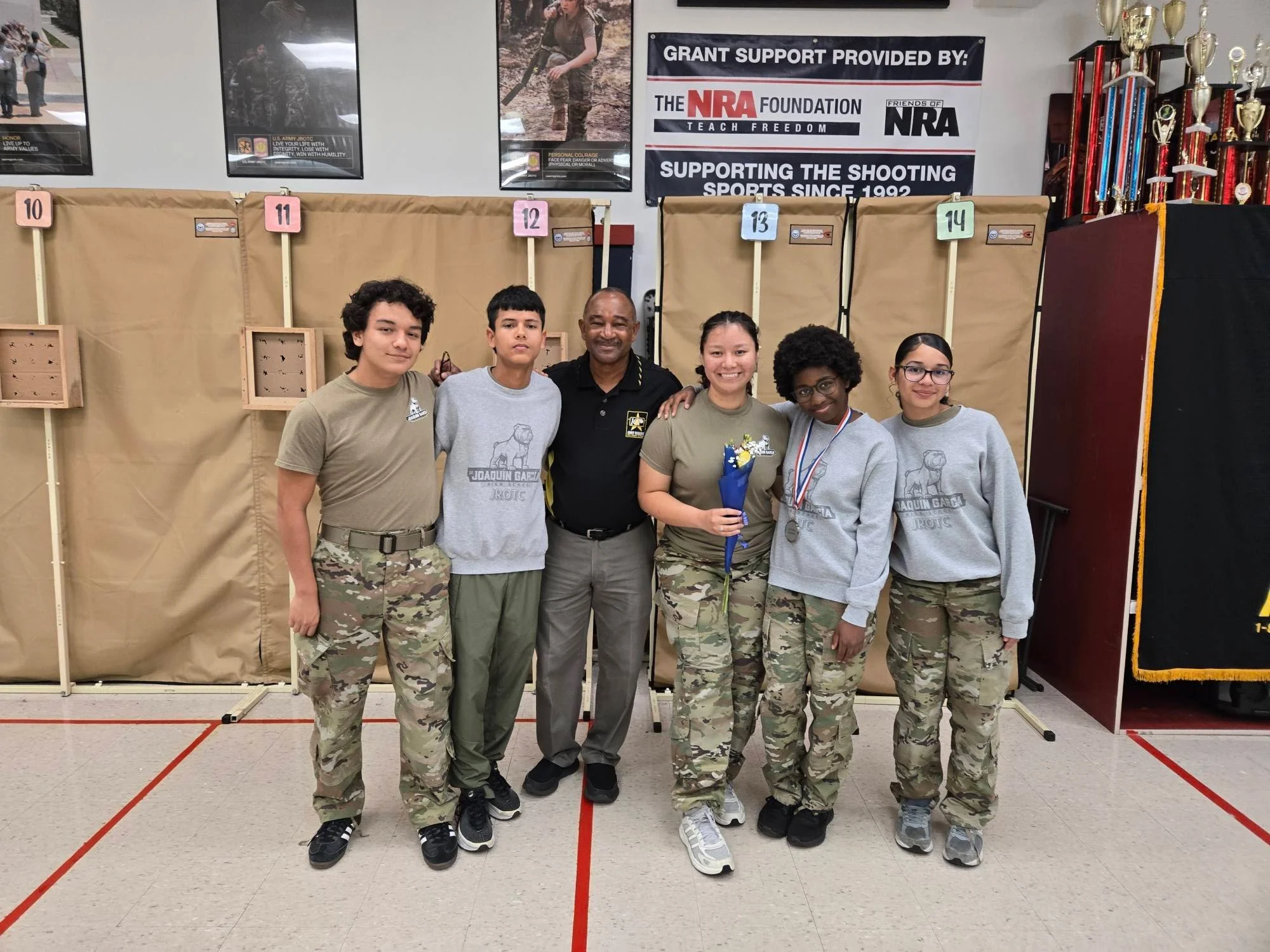 Group of six individuals, five young women and one man, inside a shooting range, with trophies and medals, posing for a photo, some wearing camouflage pants, one holding flowers, at a shooting competition.