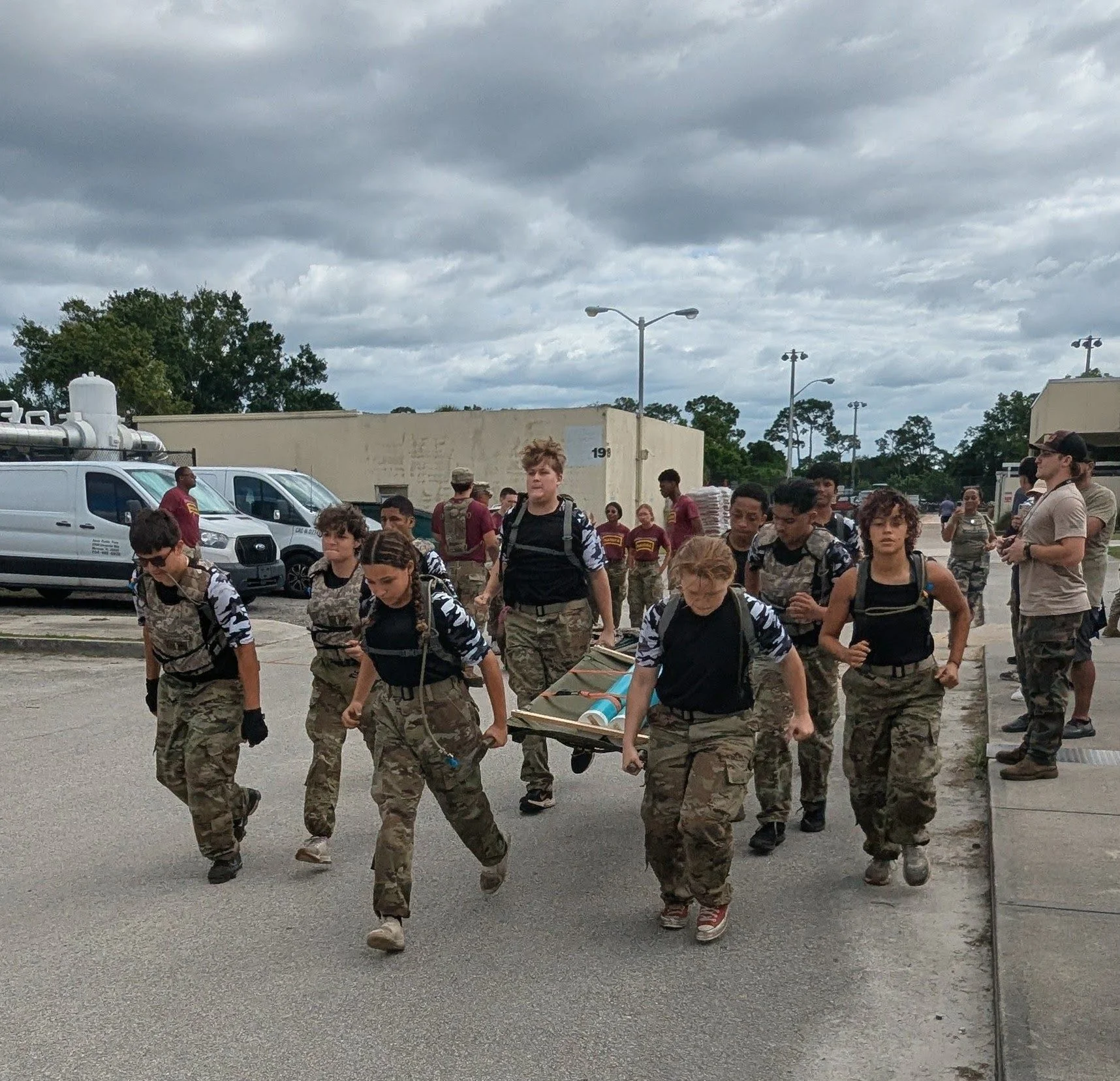 Group of young people in military-style camouflage clothing walking outdoors, carrying a stretcher with supplies, under cloudy sky.