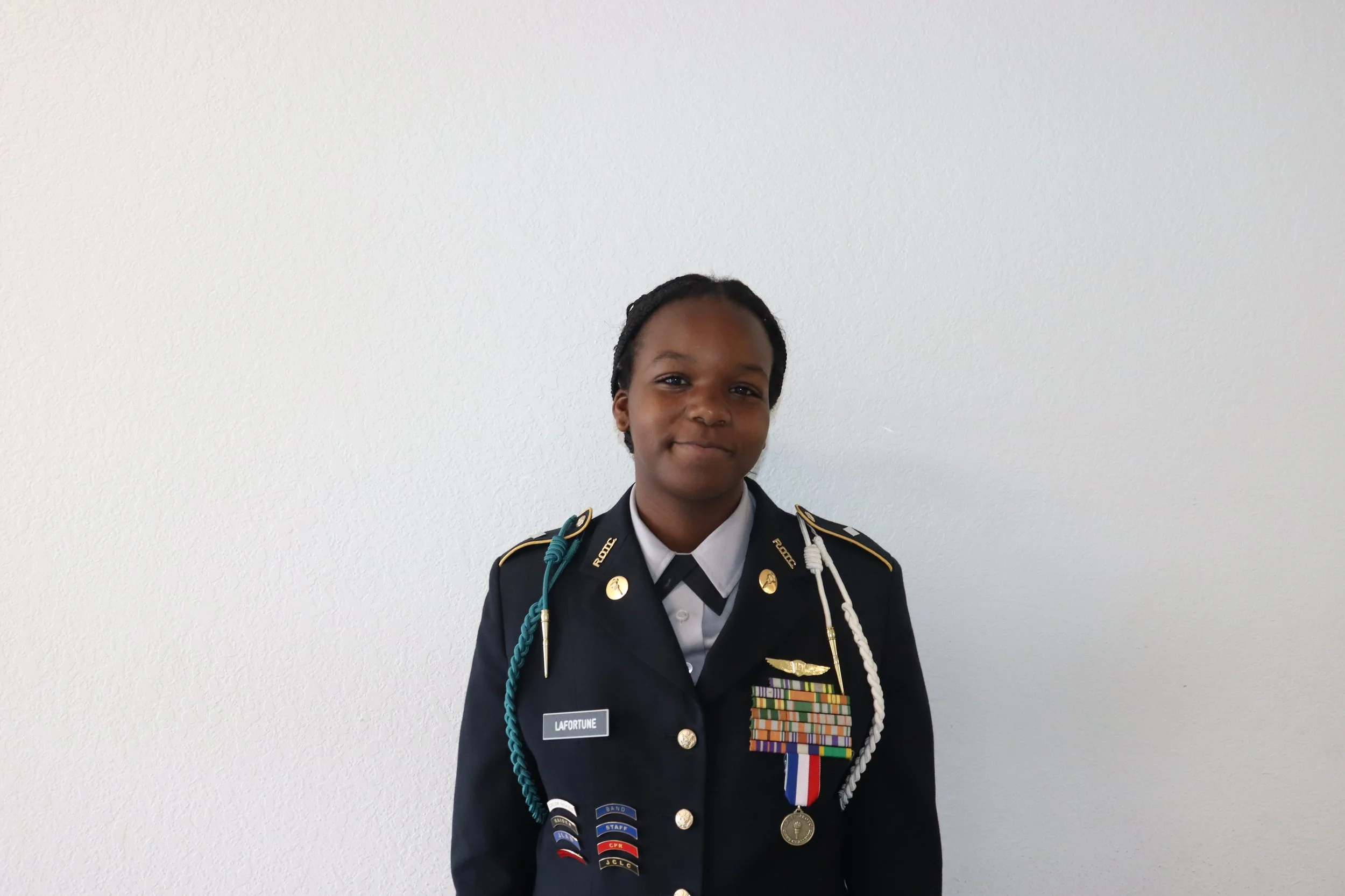 Young girl in military uniform with medals and ribbons standing against a plain white wall.