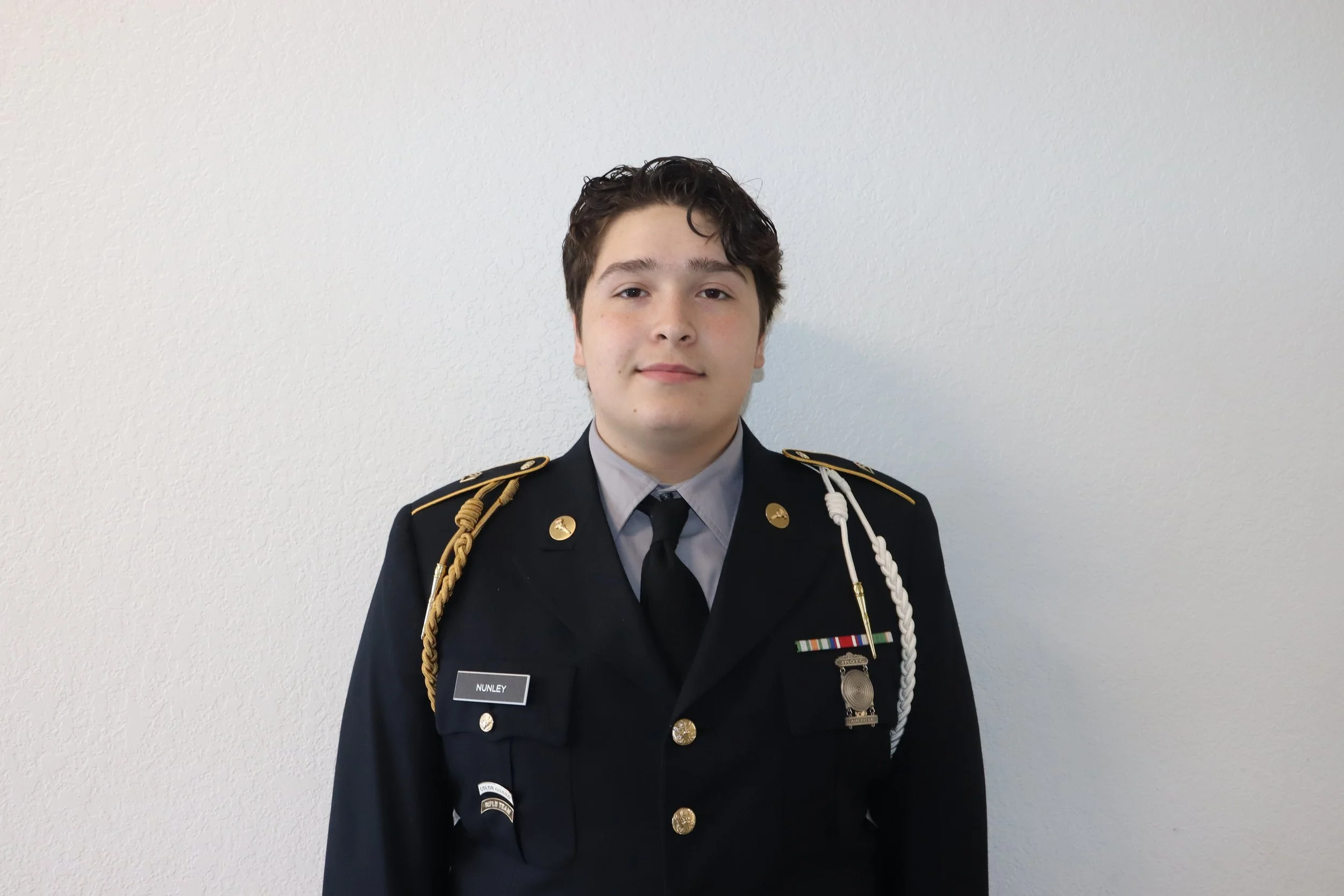 Young man in military dress uniform standing against a plain white wall.