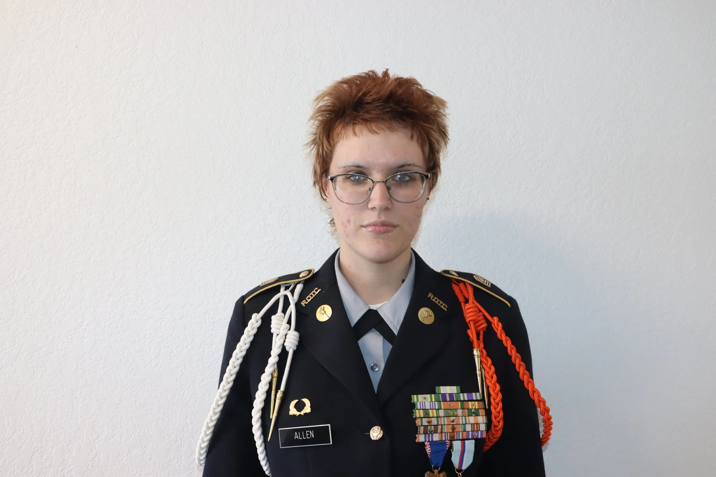 Young woman with short red hair and glasses wearing a military uniform with medals and decorations, standing against a plain white wall.