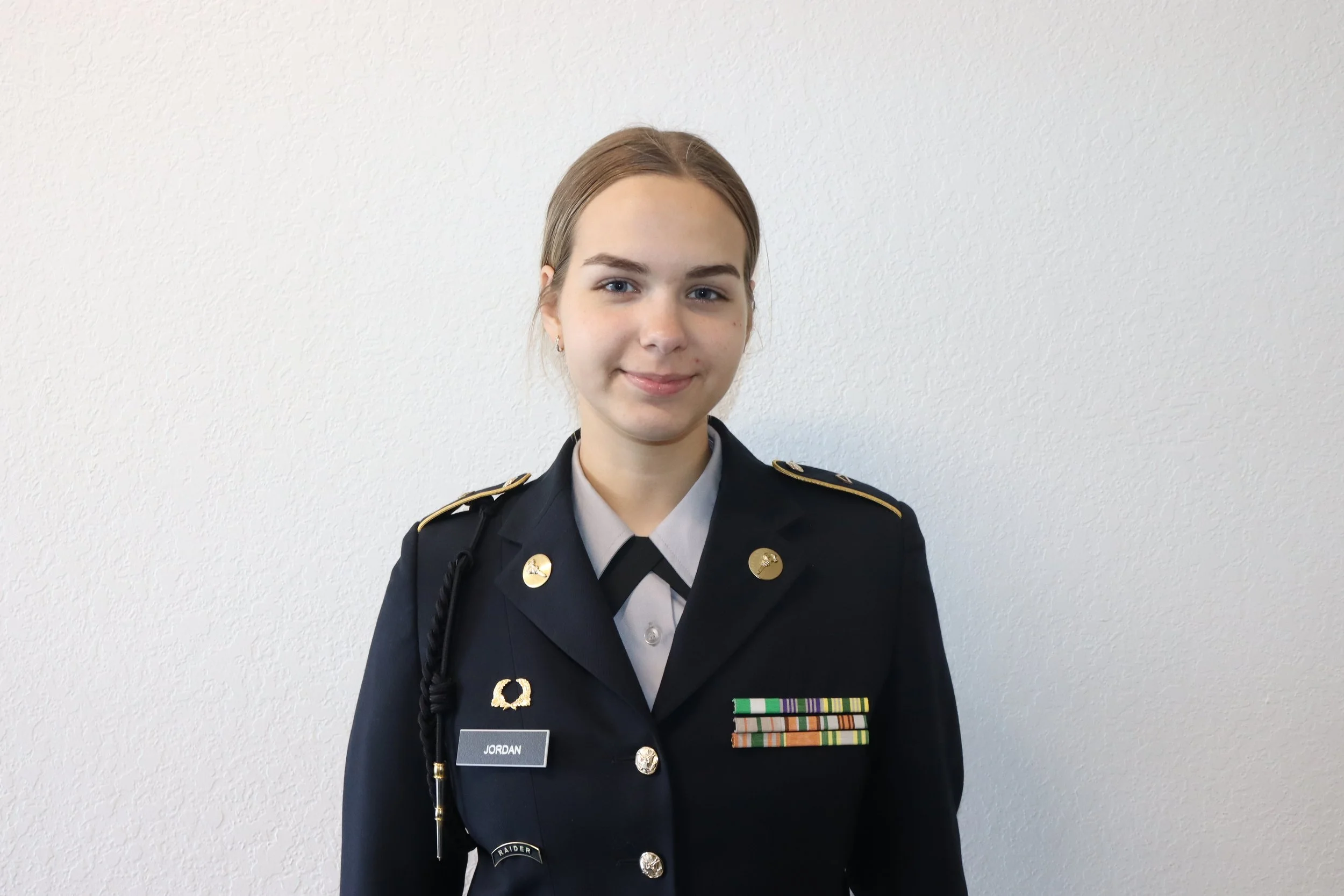 Young woman in military dress uniform standing against a plain white wall, smiling.