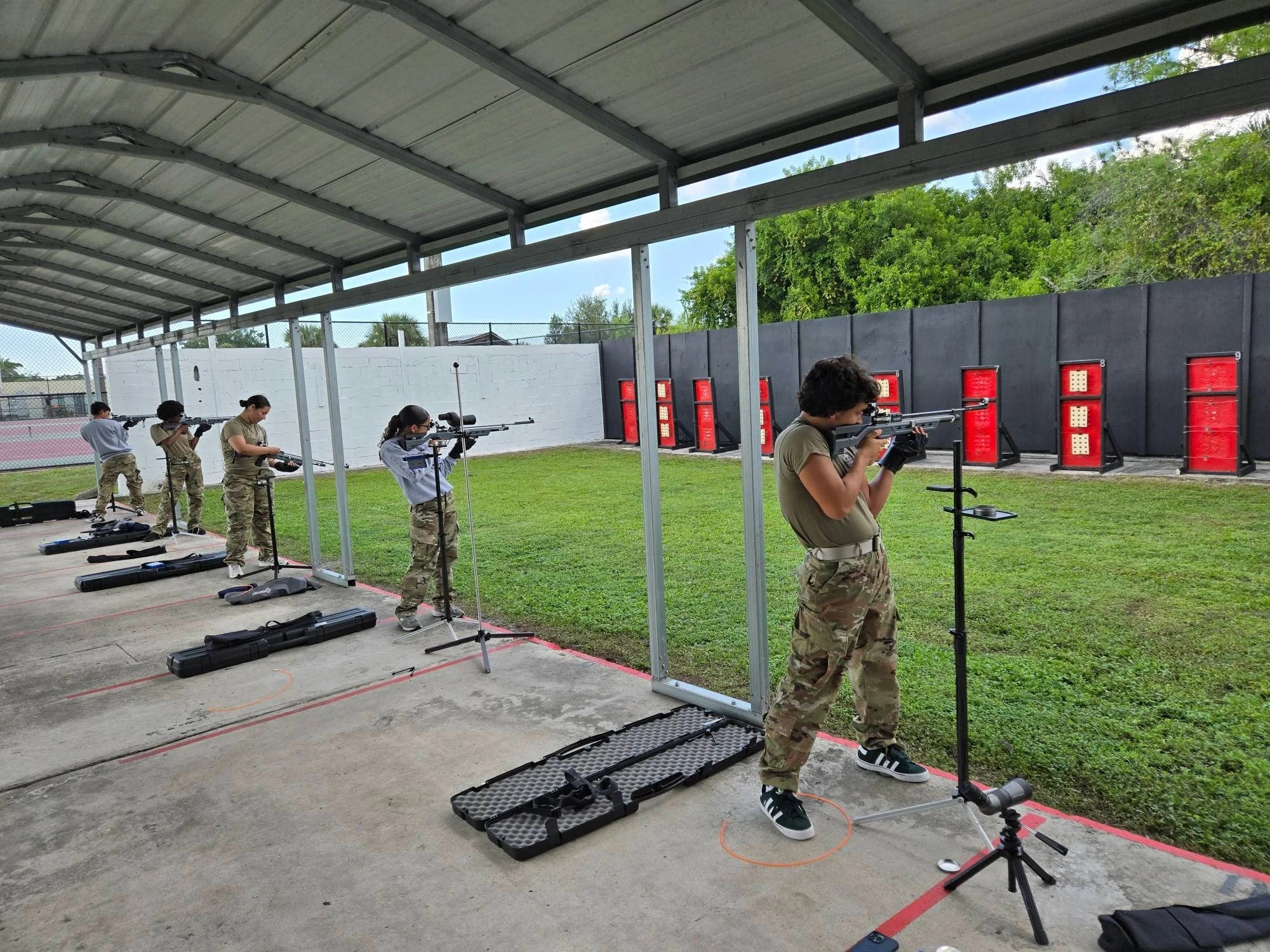Five individuals, including one woman and four men, practicing shooting at an outdoor shooting range under a covered roof. They are standing at designated spots with rifles aimed at targets downrange, with gun cases on the ground nearby and targets set up against a black wall.