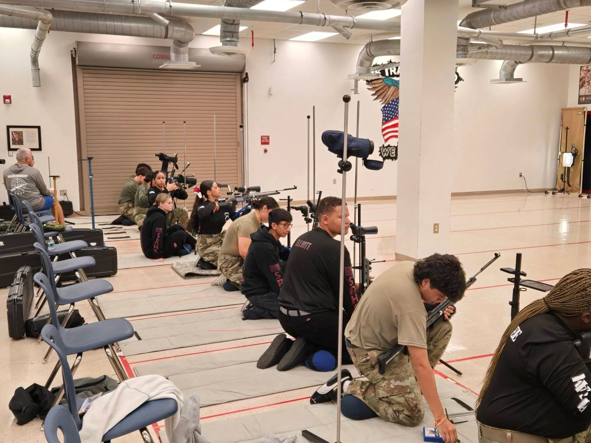 Group of people kneeling on mats, aiming rifles in an indoor shooting range.