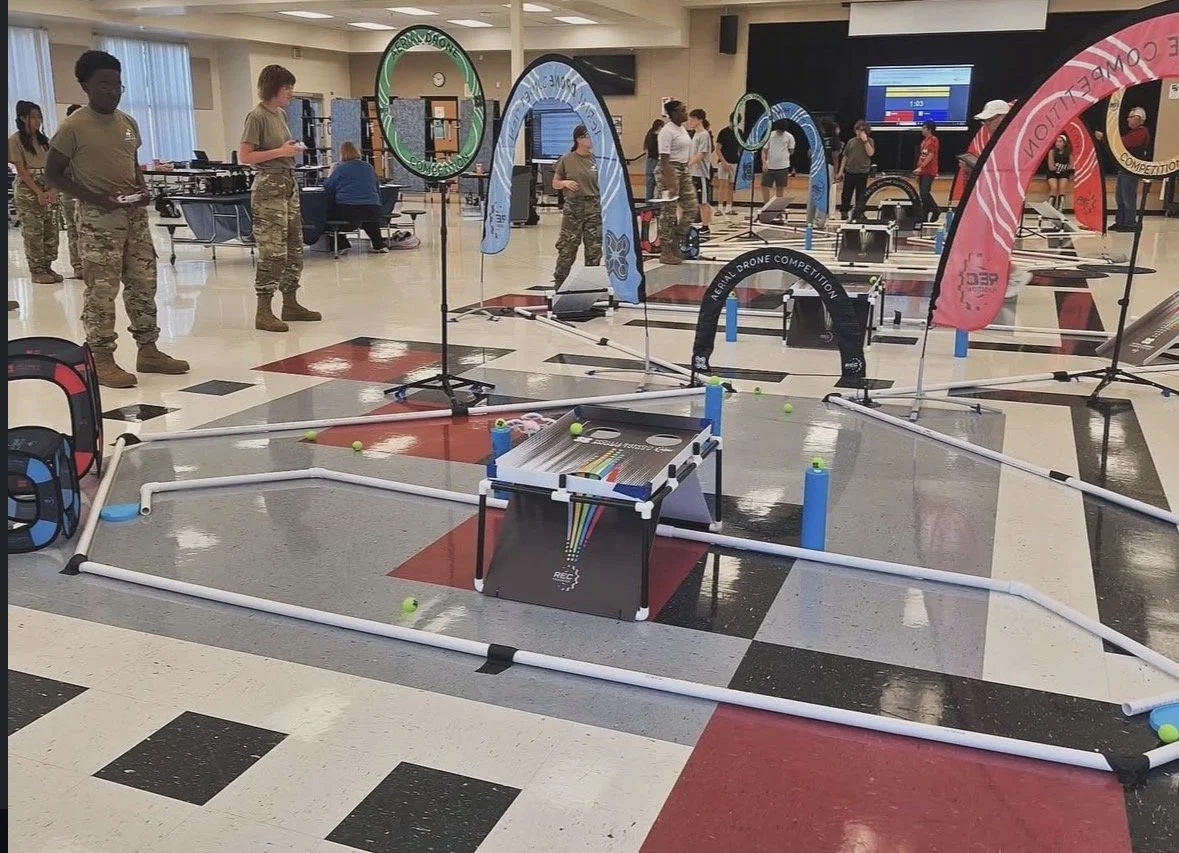 Indoor drone competition setup with banners, tables, and participants, some in military-style clothing, in a large room with a black and white checkered floor.