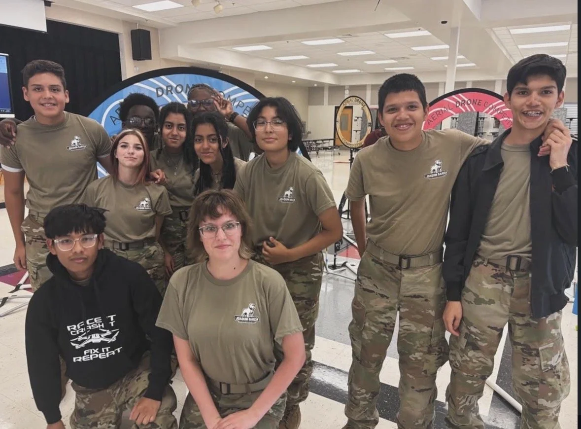 Group of students and an adult woman wearing matching military-style uniforms, standing indoors with drone display structures in the background.