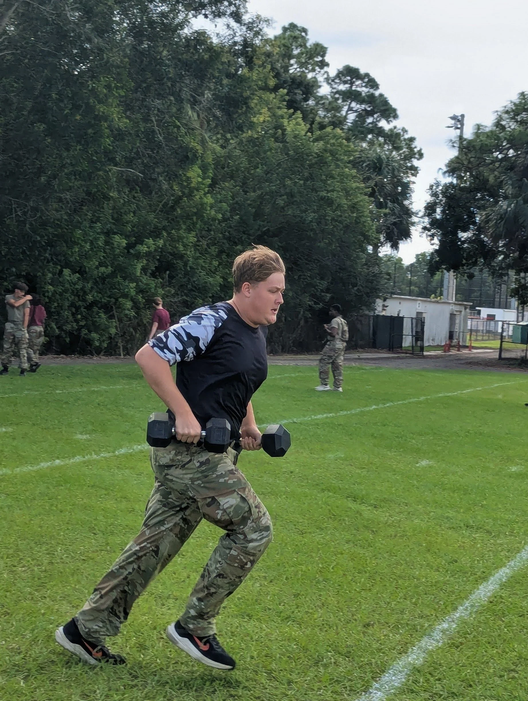 A woman in camouflage pants and a black t-shirt with camouflage sleeves exercises on a grassy field, holding dumbbells in both hands, with trees and other people in the background.