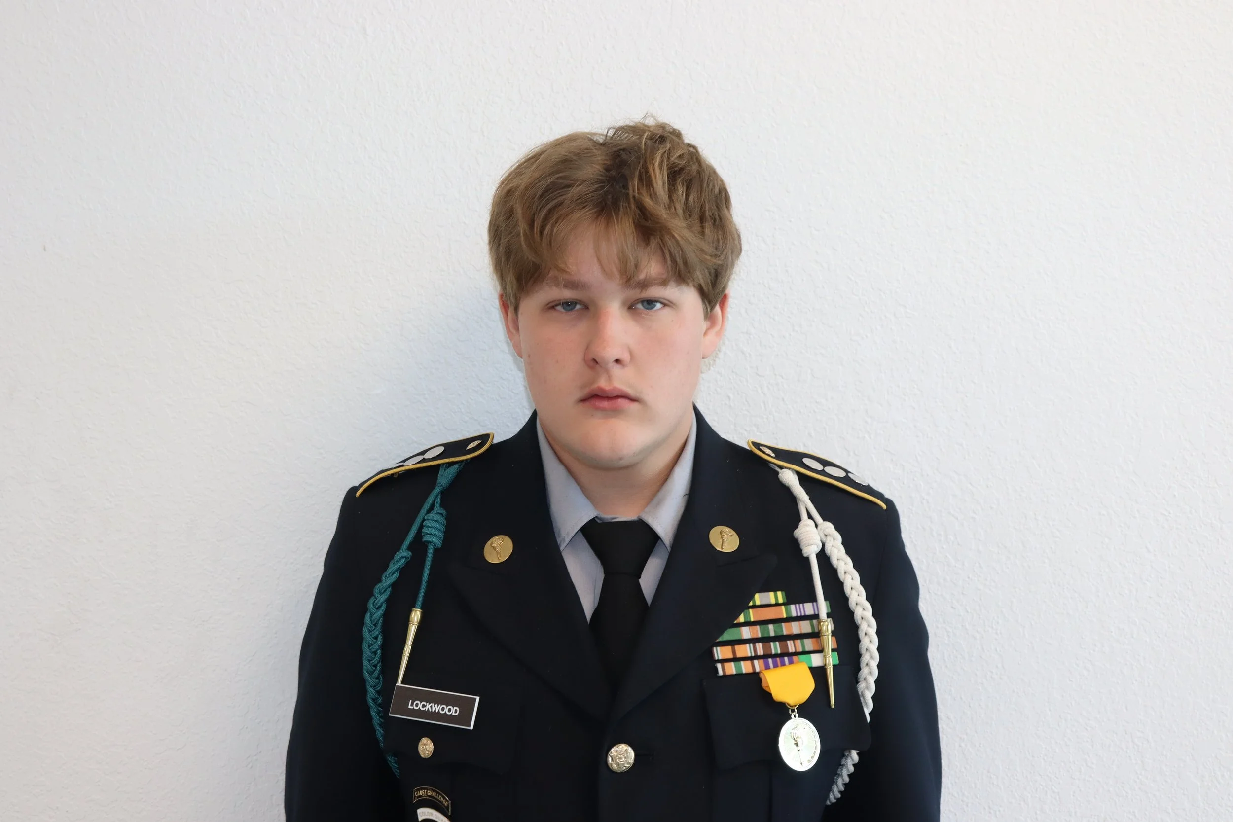 Young man in a U.S. Army dress uniform with medals and decorations standing against a plain white wall.
