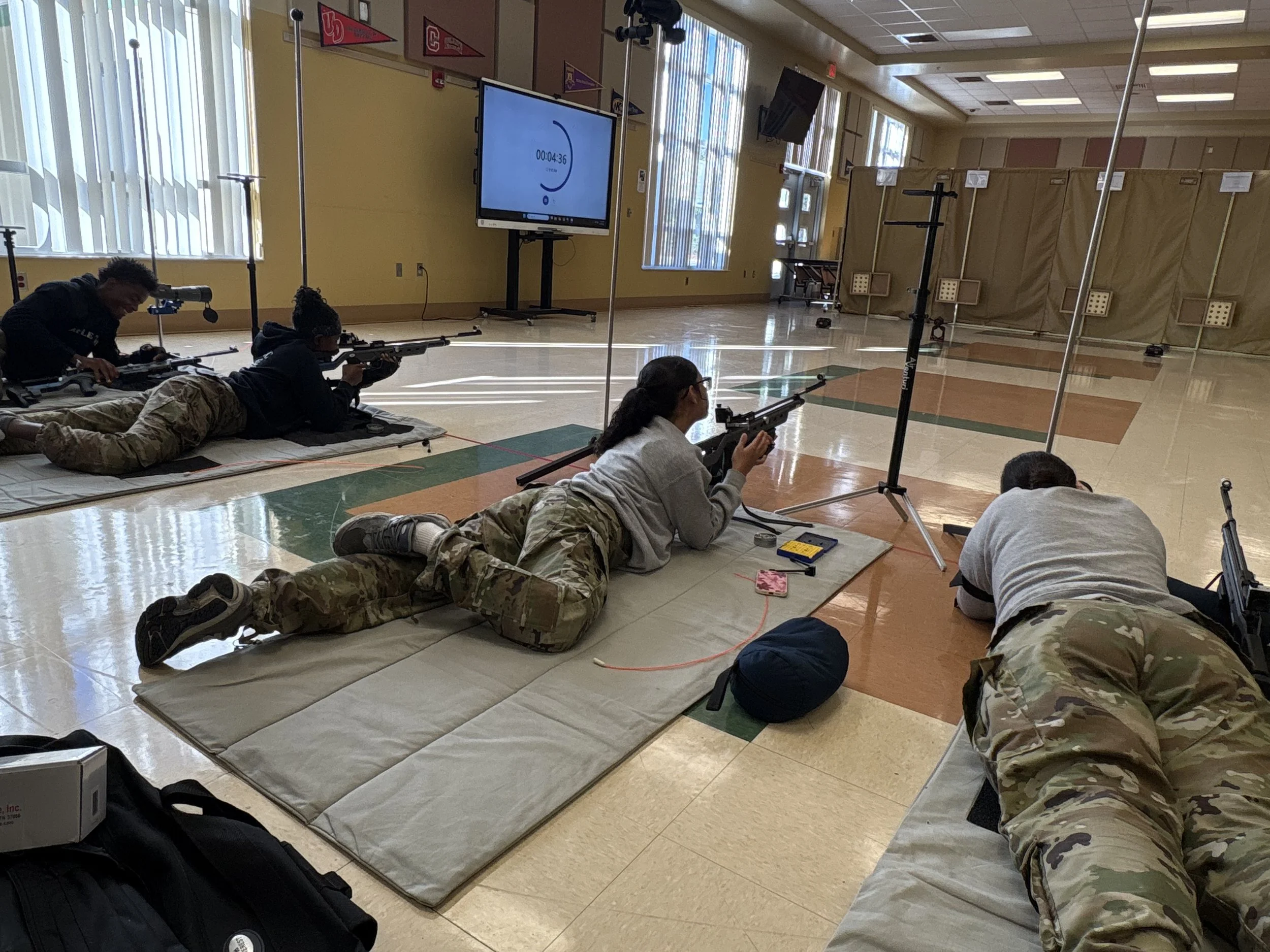 Four individuals lying on mats in a gymnasium, aiming rifles at targets in an indoor shooting range. They are in prone position, with one using a spotting scope, and safety gear visible.