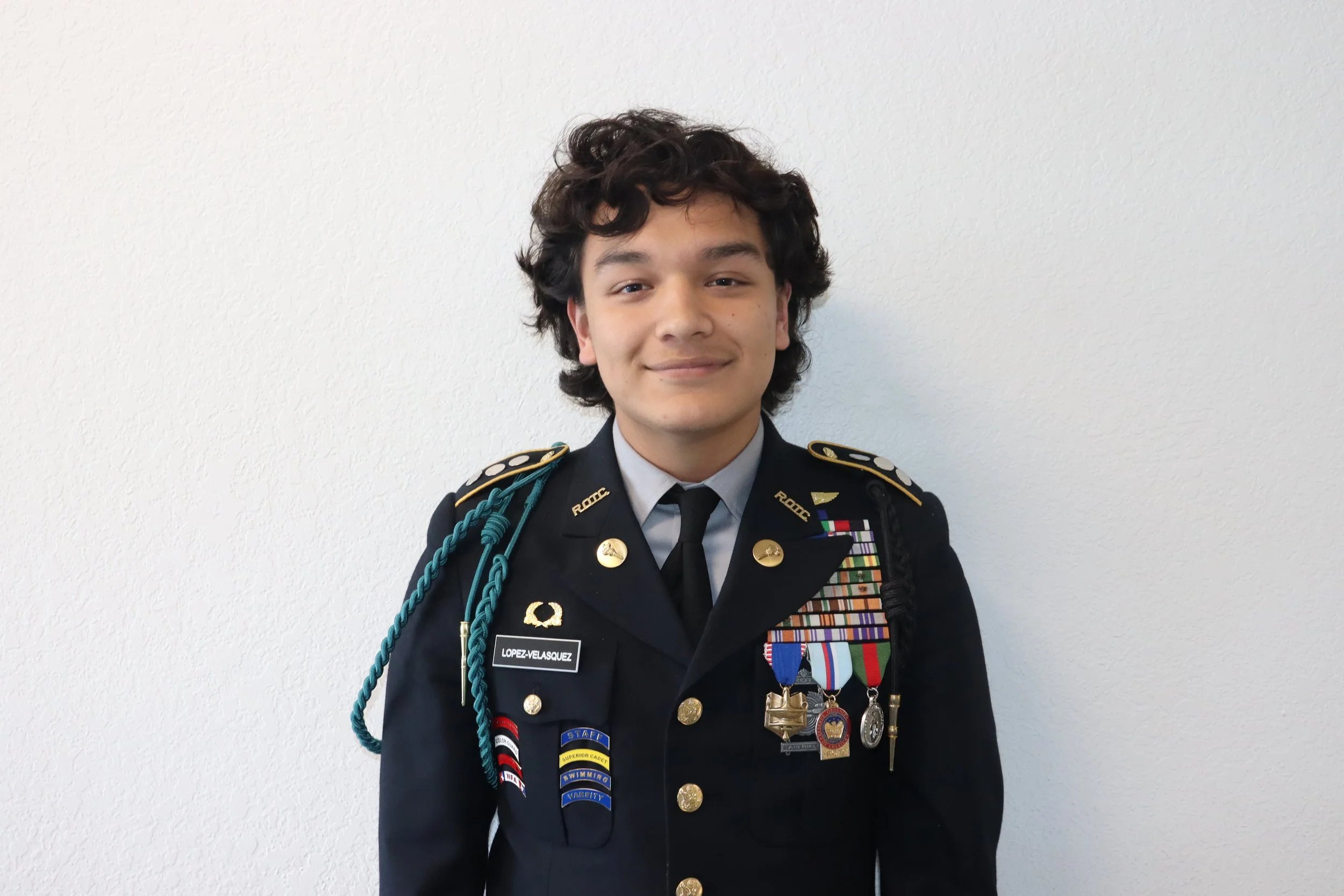 Young man in military uniform with medals and patches, standing against a white wall.