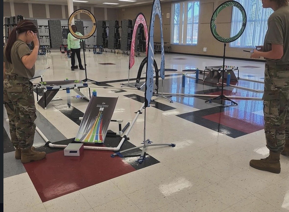 Indoor drone competition setup with two women in military-style clothing, one on each side, and several hoops and obstacles in a spacious room with large windows and shelves in the background.