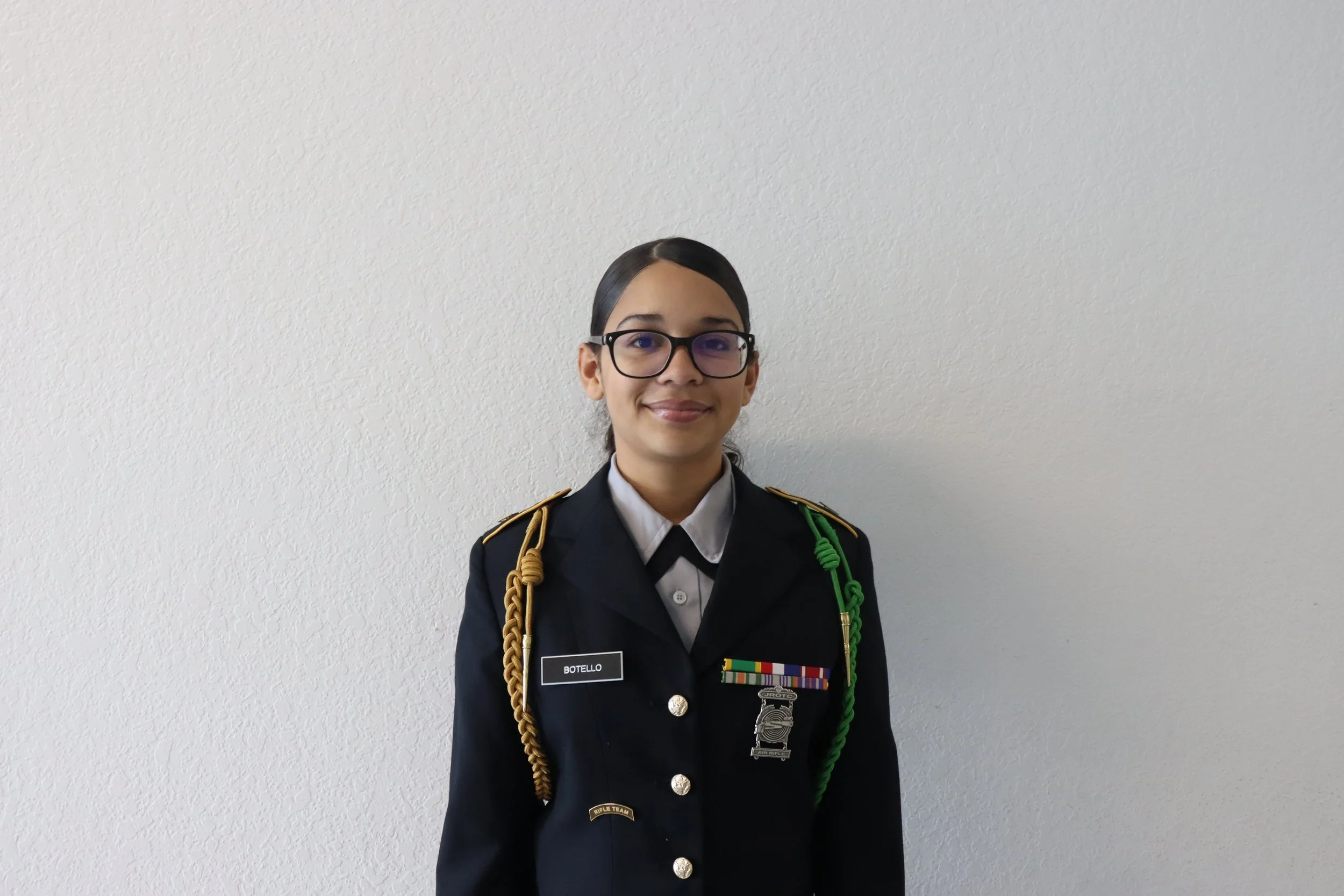 Young woman in military uniform with glasses, standing against a light-colored wall.