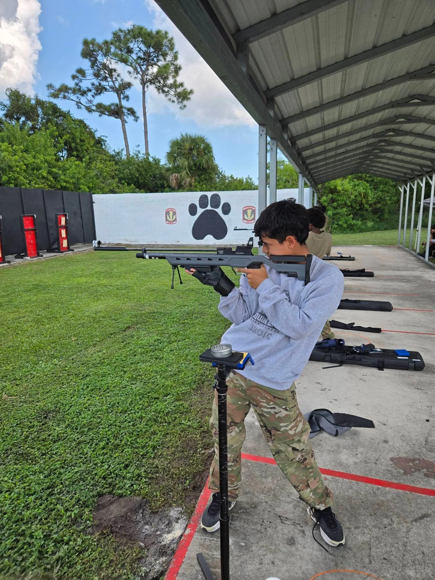 A young man at a shooting range aiming a rifle, with targets and a decorative wall featuring a large black paw print in the background.