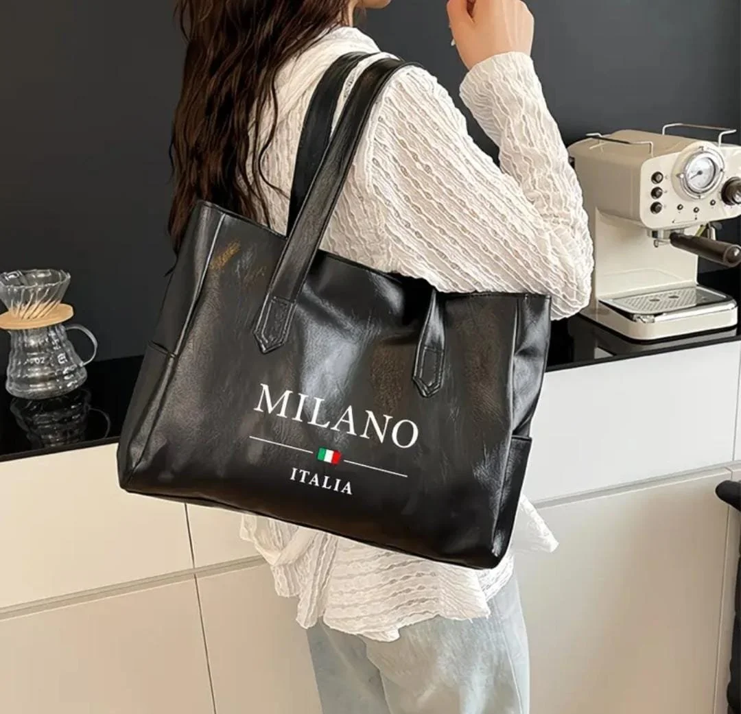 A person with dark hair carrying a large black tote bag with the words 'Milano Italia' and an Italian flag printed on it. They are standing in a modern kitchen or coffee station near a white espresso machine.