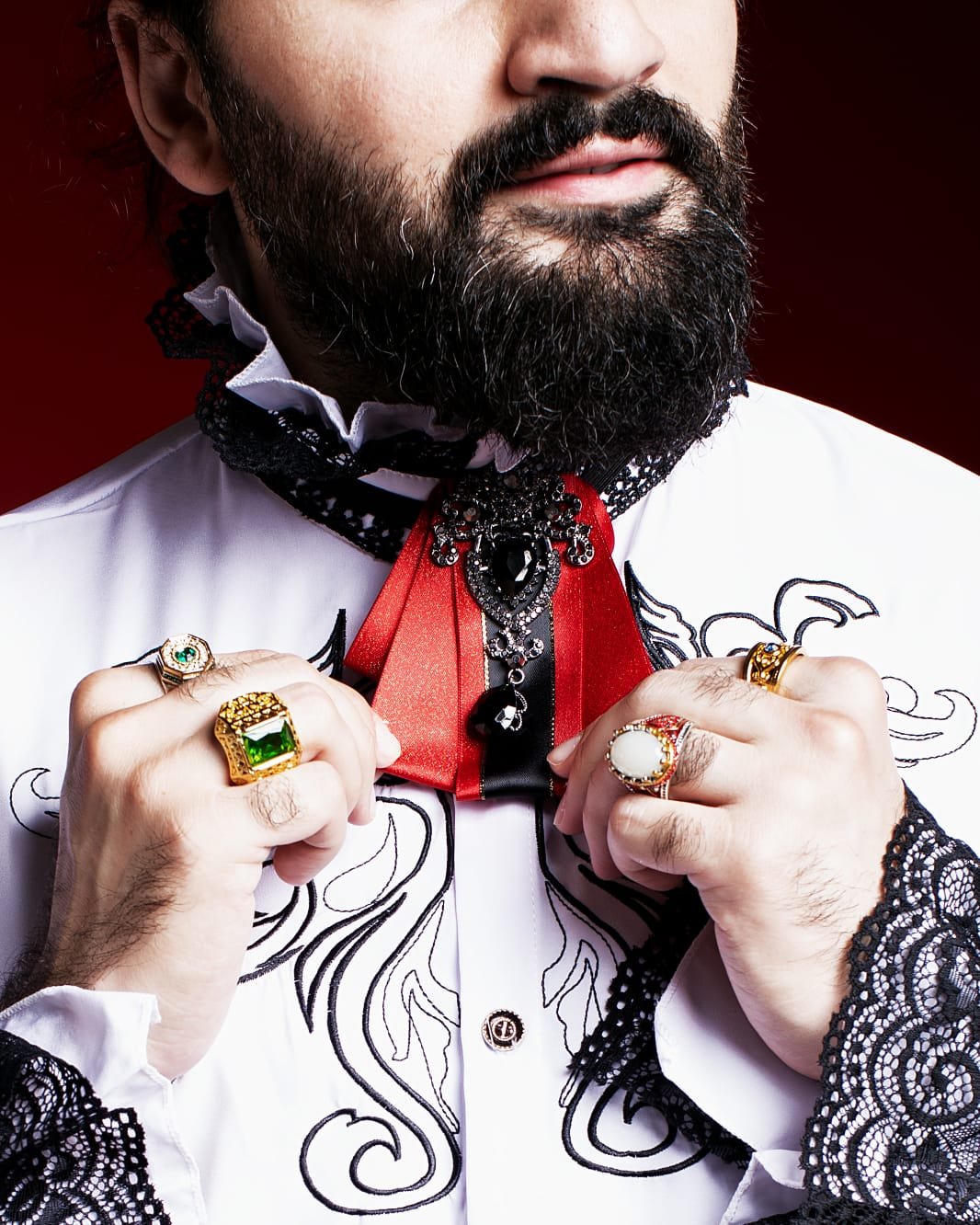 Close-up of a man with a beard, wearing ornate jewelry including rings and a decorative collar with red and black accents, on a white embroidered shirt with black lace details.