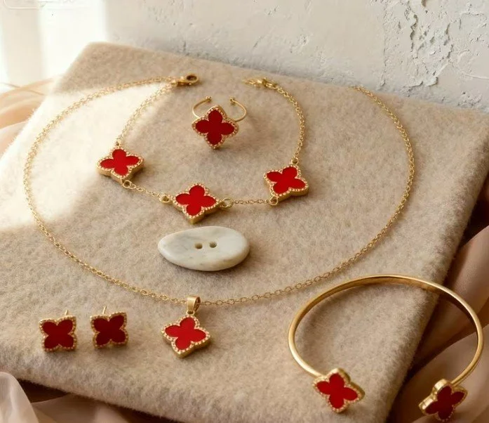 Gold jewelry set with red quatrefoil designs, including a necklace, bracelet, earrings, ring, and a white button-shaped accessory, displayed on a beige fabric surface.
