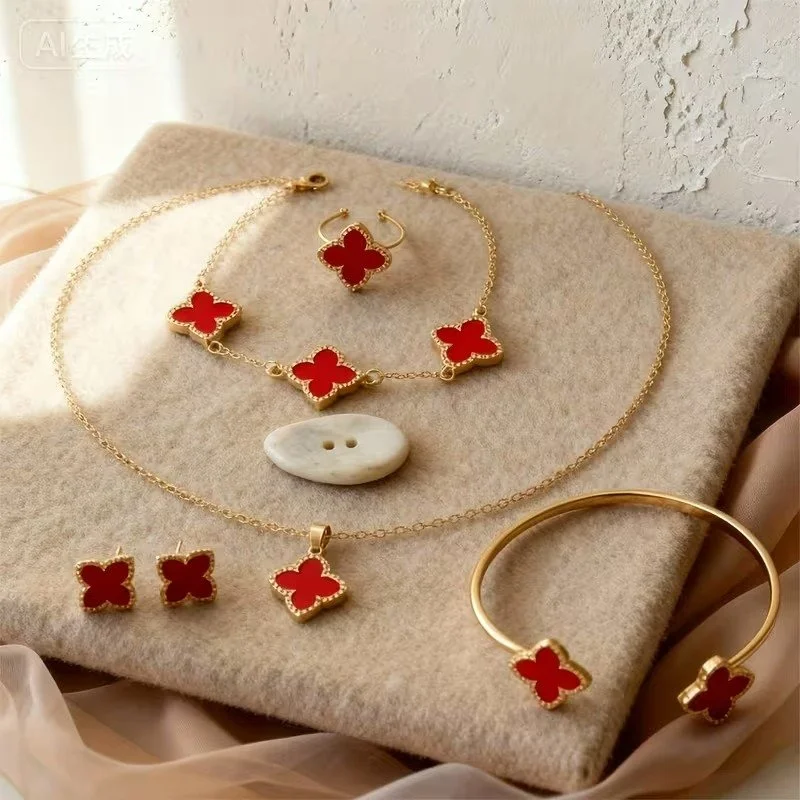Gold jewelry set with red clover-shaped stones, including a necklace, bracelet, earrings, a ring, and a collar pin, displayed on a beige fabric background.