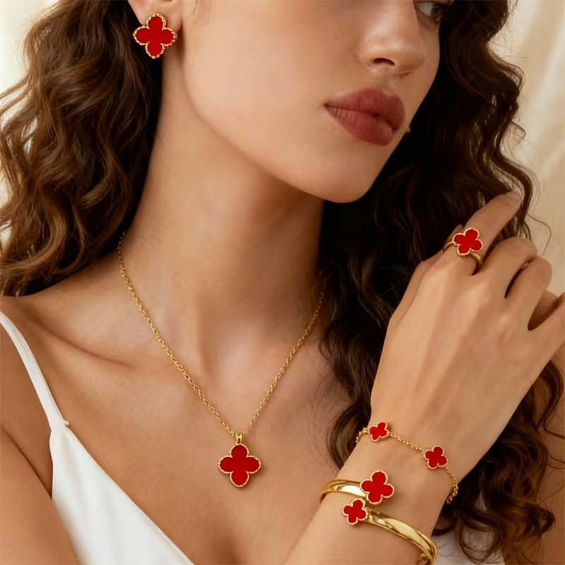 Close-up of a woman wearing jewelry with red four-leaf clover designs, including earrings, necklace, ring, bracelet, and two gold bangles.
