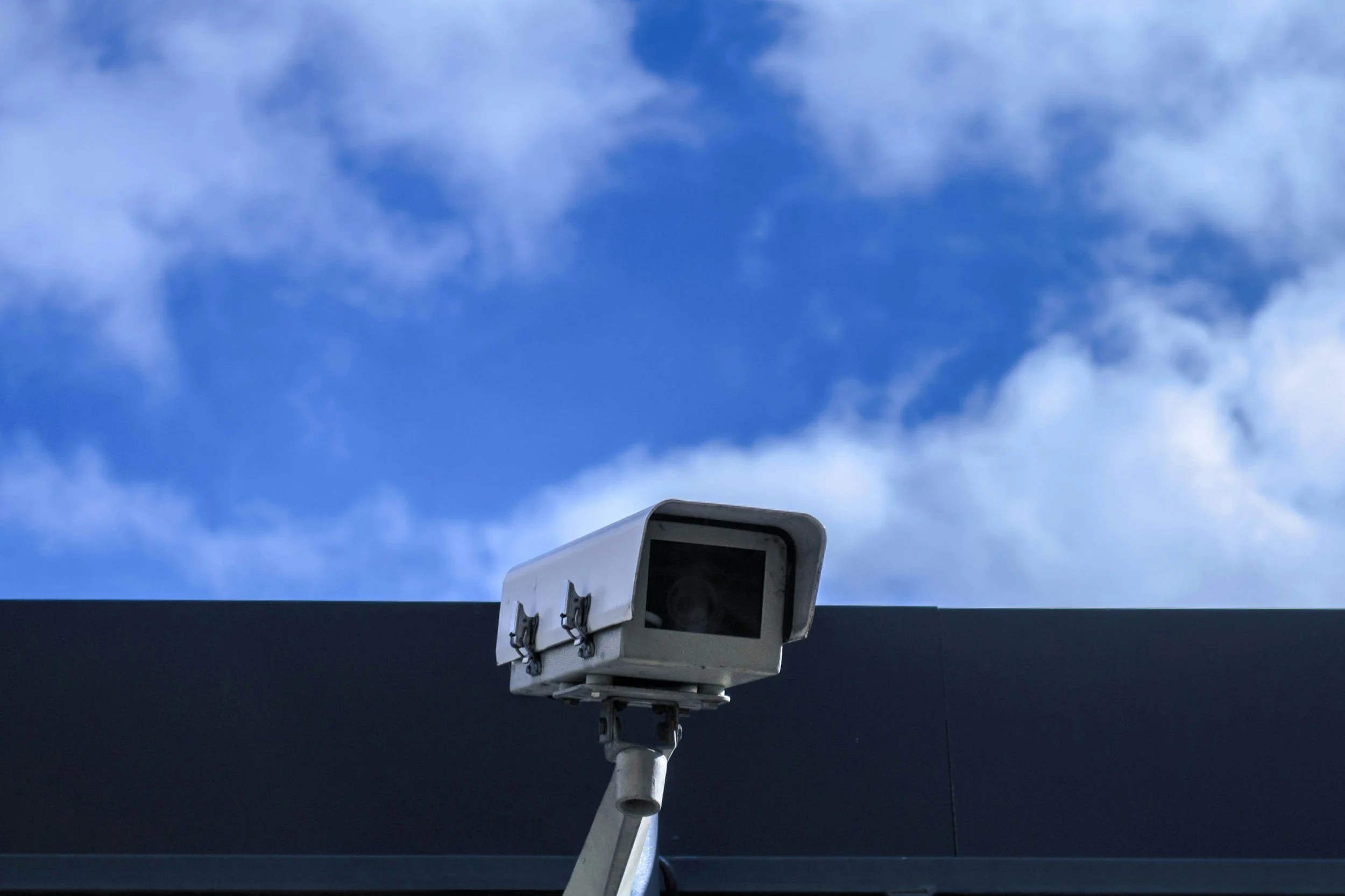 Security camera mounted on a building facing upward with a partly cloudy blue sky in the background.