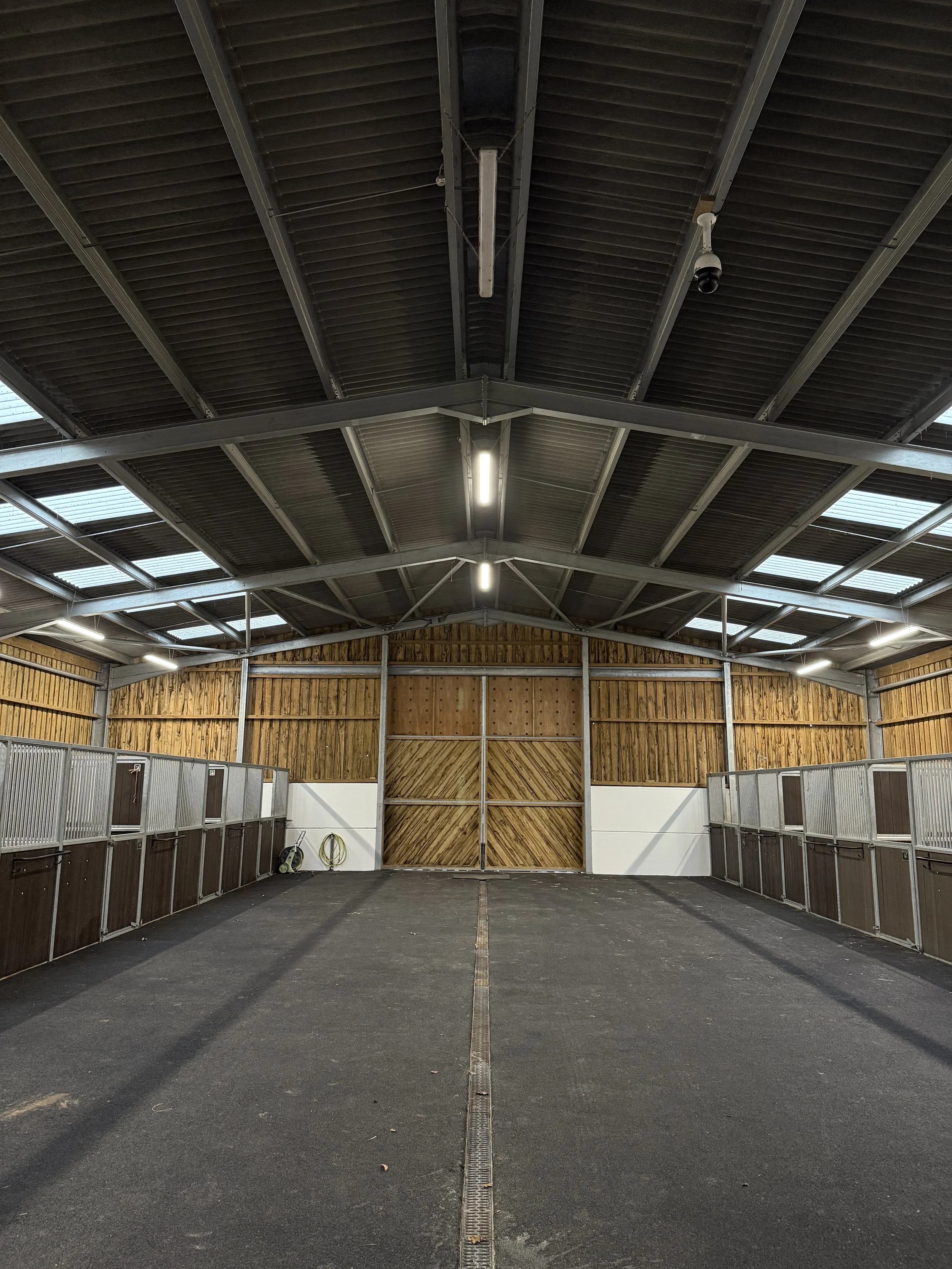 Empty horse stable with wooden and metal stalls, concrete floor, and a large wooden sliding door at the back, inside a spacious barn with metal roof and skylights.