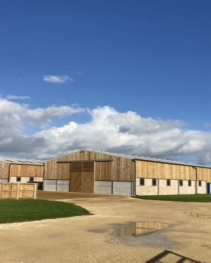 A large wooden barn with a clear blue sky and scattered clouds in the background, surrounded by a gravel driveway and some green grass.
