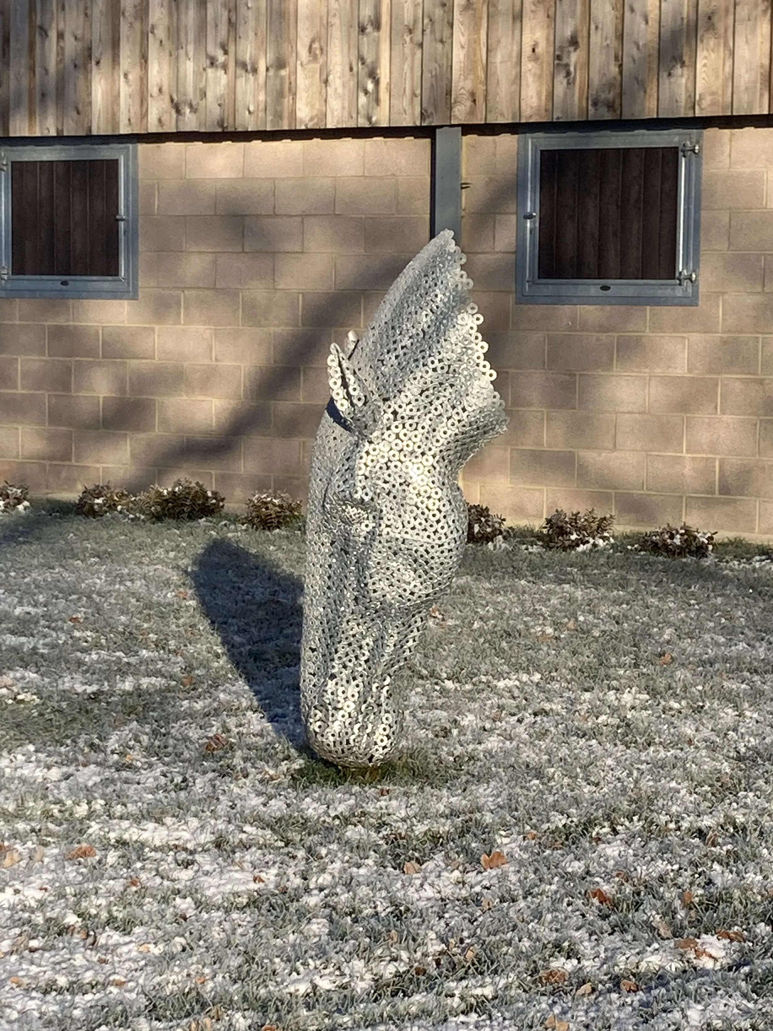 A metallic owl sculpture made of washers, standing outdoors on a frost-covered grass area, with a wooden and stone building in the background.