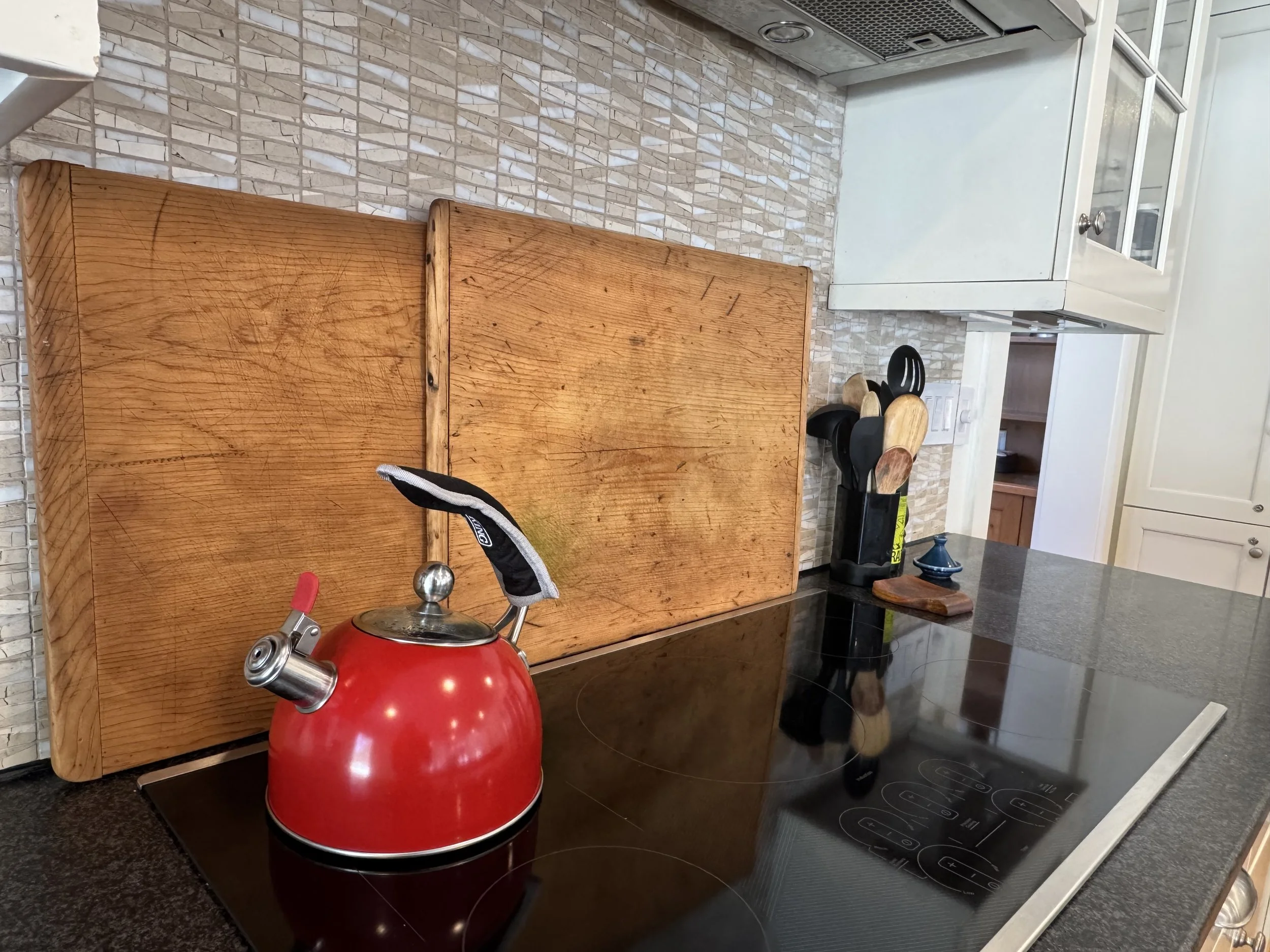 A red kettle on a black electric stove with a wooden backsplash and kitchen utensils in a holder on the counter.