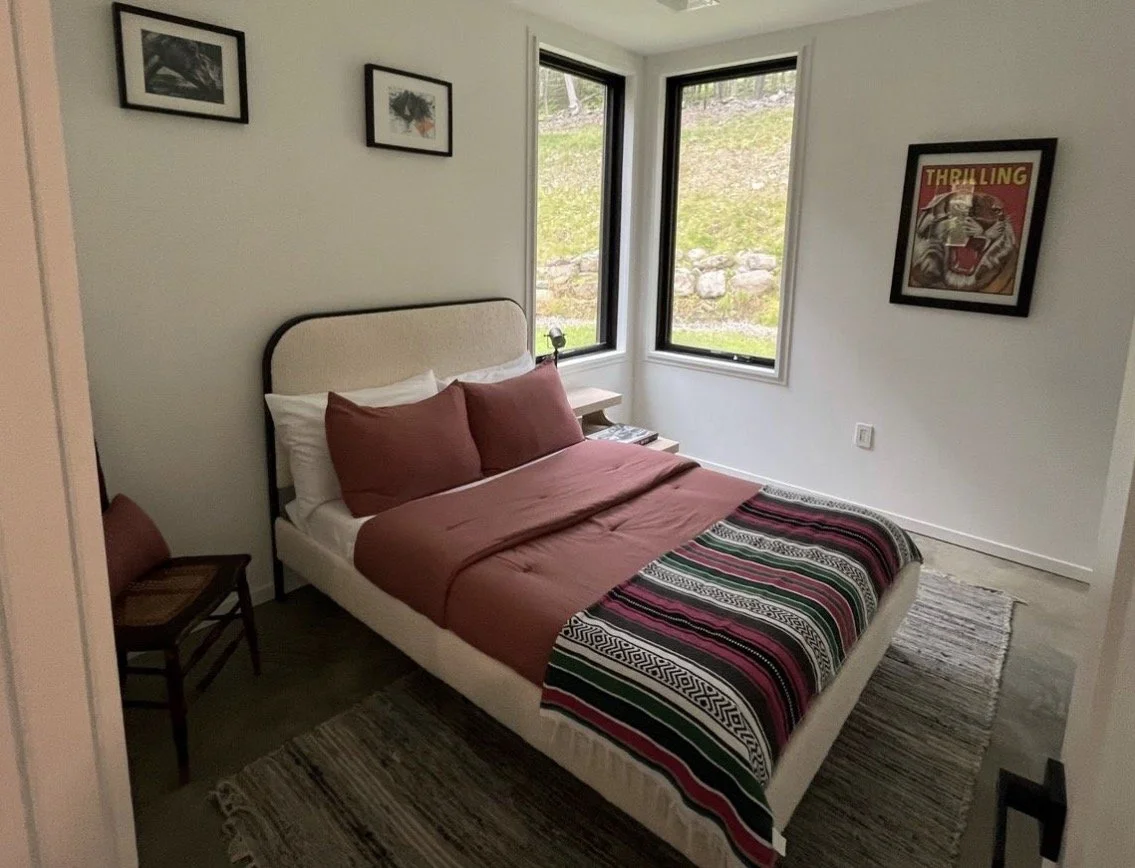 A bedroom with a bed, two pillows, a blanket, a wooden chair, and framed pictures on the wall, near two windows showing a grassy outdoor area.