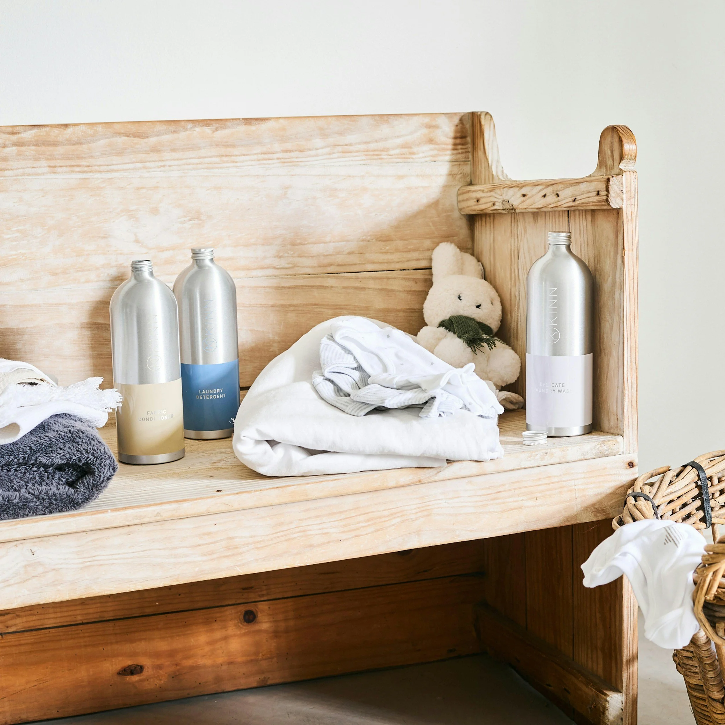 A wooden bench with laundry detergent bottles, folded clothes, and a stuffed bunny plush toy on it, next to a woven basket filled with more laundry.