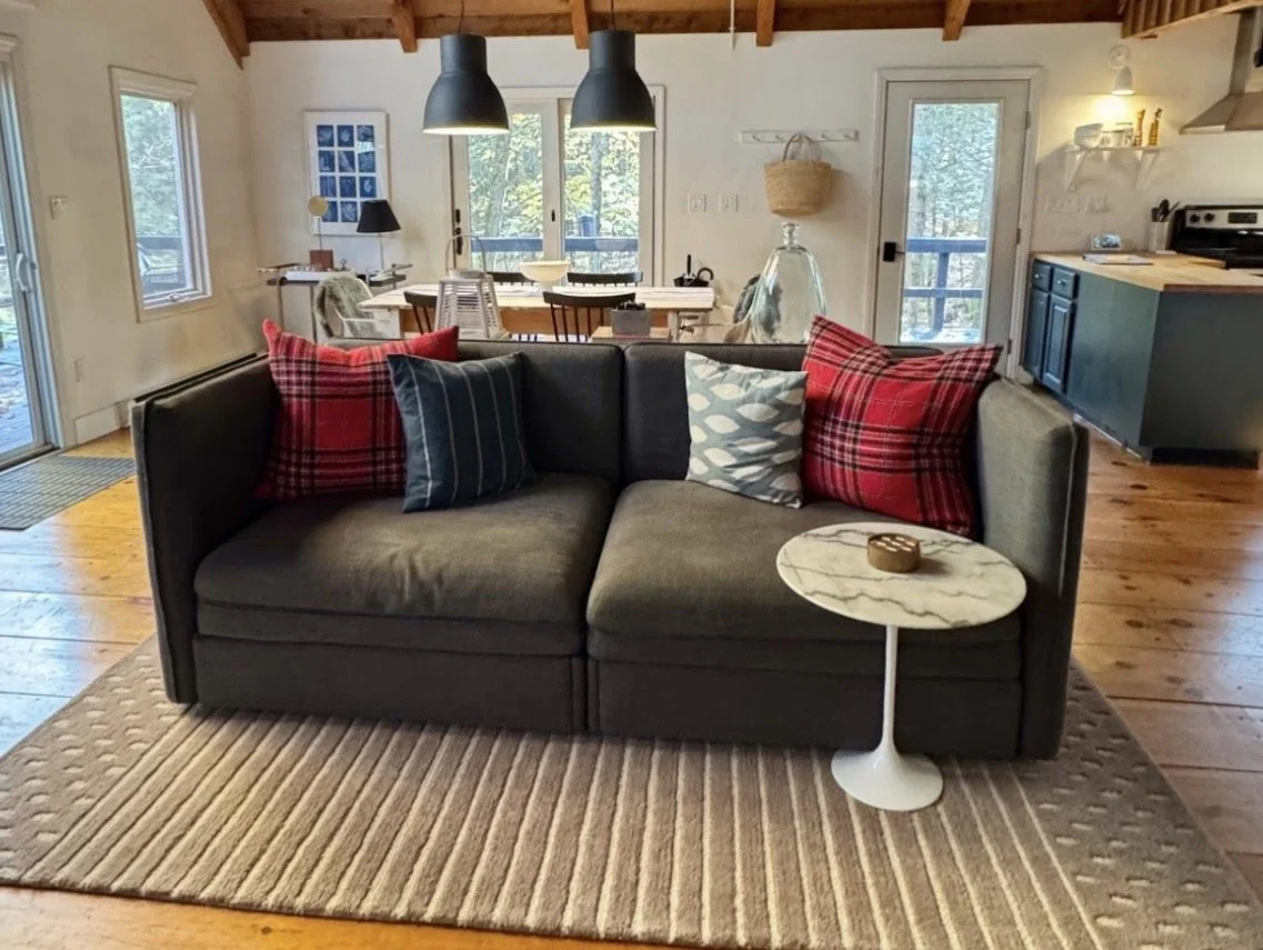 A living room with a dark sofa with multiple pillows, a small round marble side table, a striped rug, and a wooden dining table with chairs in the background. There are large windows, a door, and various decorations including a woven basket and lamps