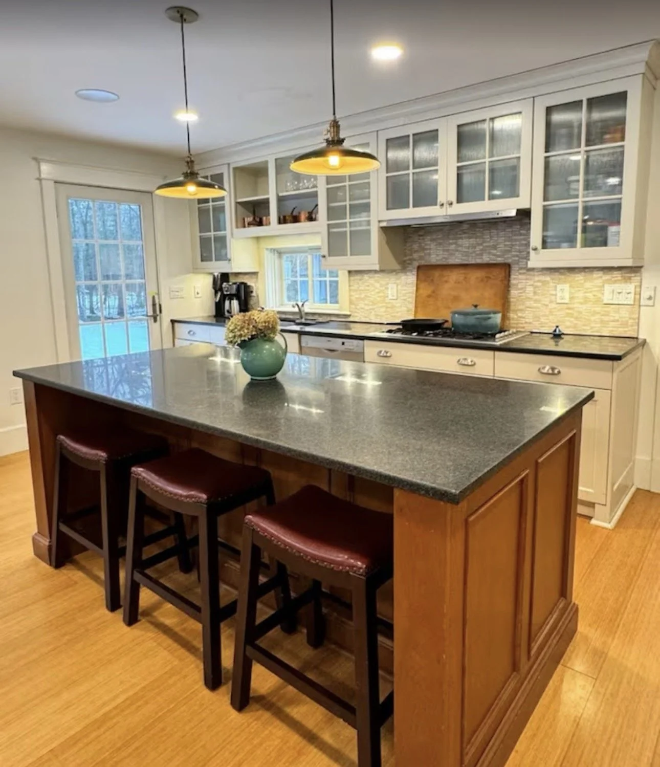 A kitchen with a large island, bar stools, white cabinets with glass doors, a black countertop, and a backsplash with beige tiles. There are pendant lights hanging from the ceiling and a door leading outside.