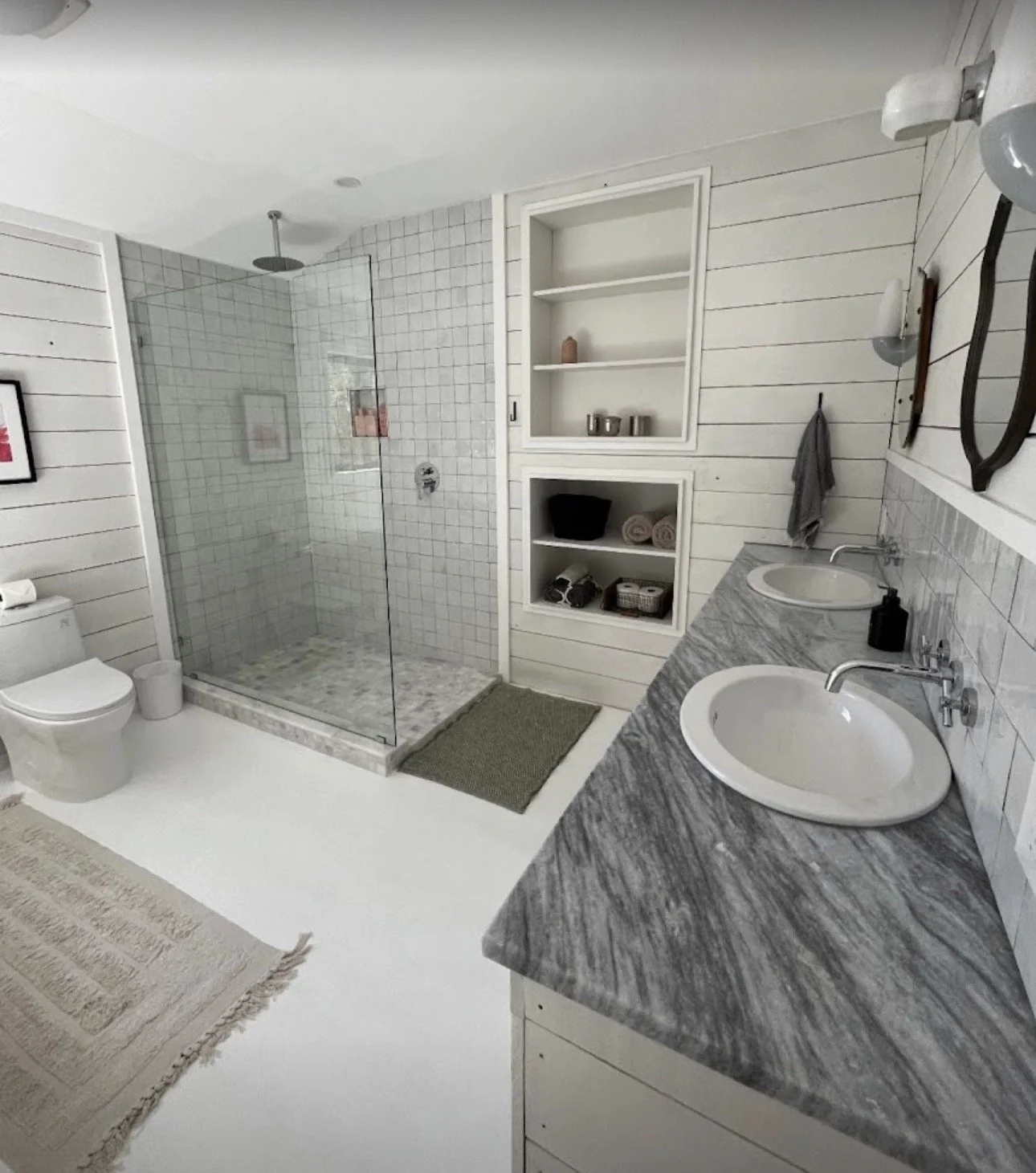 A modern bathroom with a double sink marble countertop, open shelves, a toilet, and a glass-enclosed walk-in shower with white tile walls.