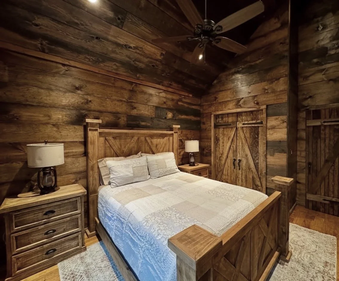 A rustic bedroom with wooden walls, a bed with a wooden headboard and matching side tables, and two lamps on either side of the bed.
