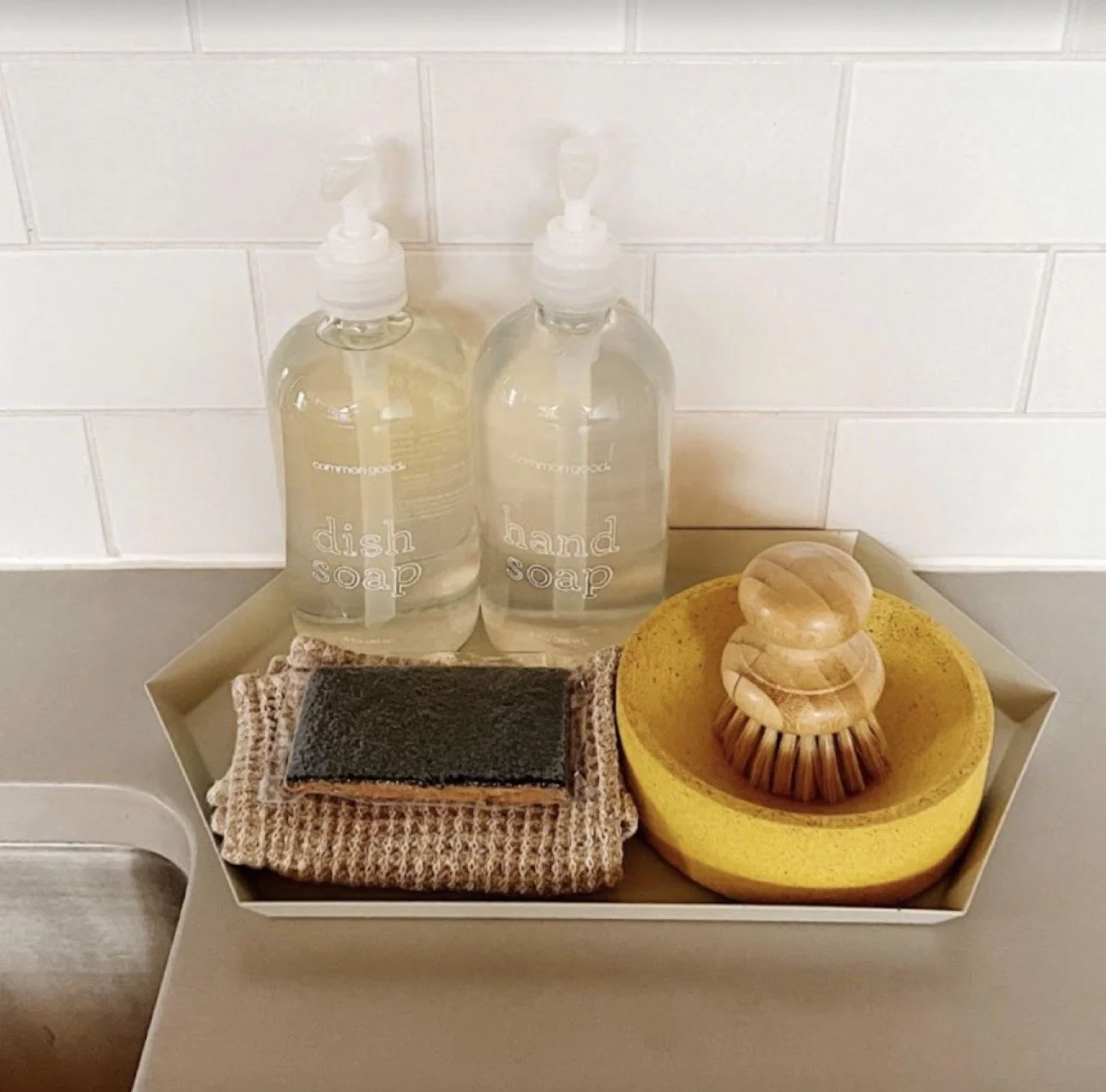 A tray with two bottles of dish soap and hand soap, a black scrub brush, a beige textured cloth, and a yellow scrub sponge, all placed on a gray surface in front of a white tiled wall.