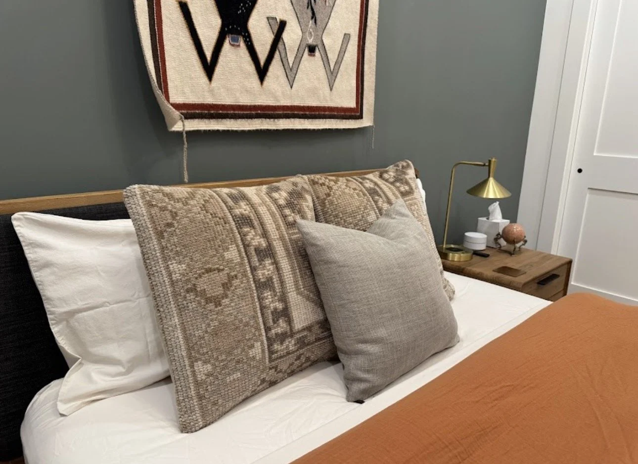 A neatly made bed with white sheets, decorative pillows including a large patterned one and a smaller gray one. Next to the bed, a wooden nightstand holds a gold-colored lamp, a box of tissues, and decorative items. A textured wall hanging with geometric shapes is above the bed, and part of a white closet door is visible on the right.