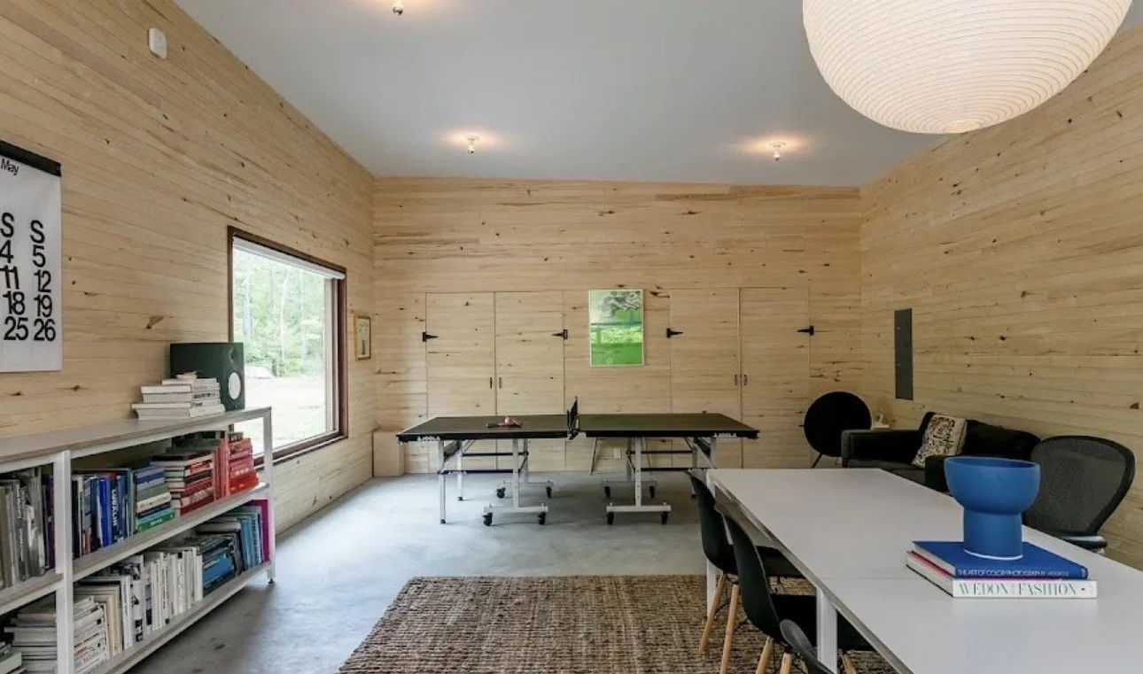 Room with wooden walls, a large window, ping pong table, bookshelf, white table with black chairs, black couch, patterned pillow, blue vase, book, and hanging paper lantern