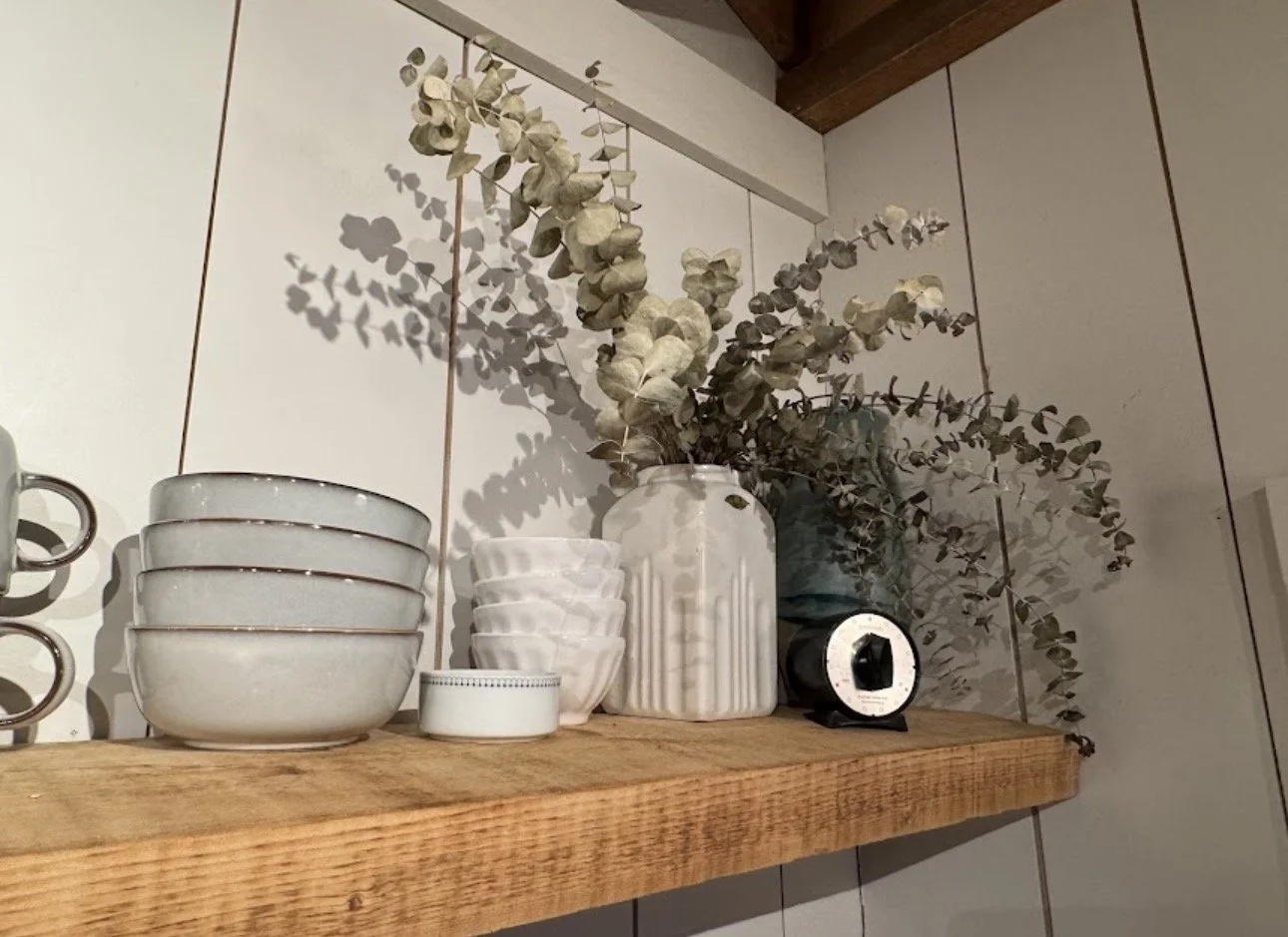 Wooden shelf with white ceramic bowls, vases, dried eucalyptus, a small black timer, and a dark bottle against a tiled wall.