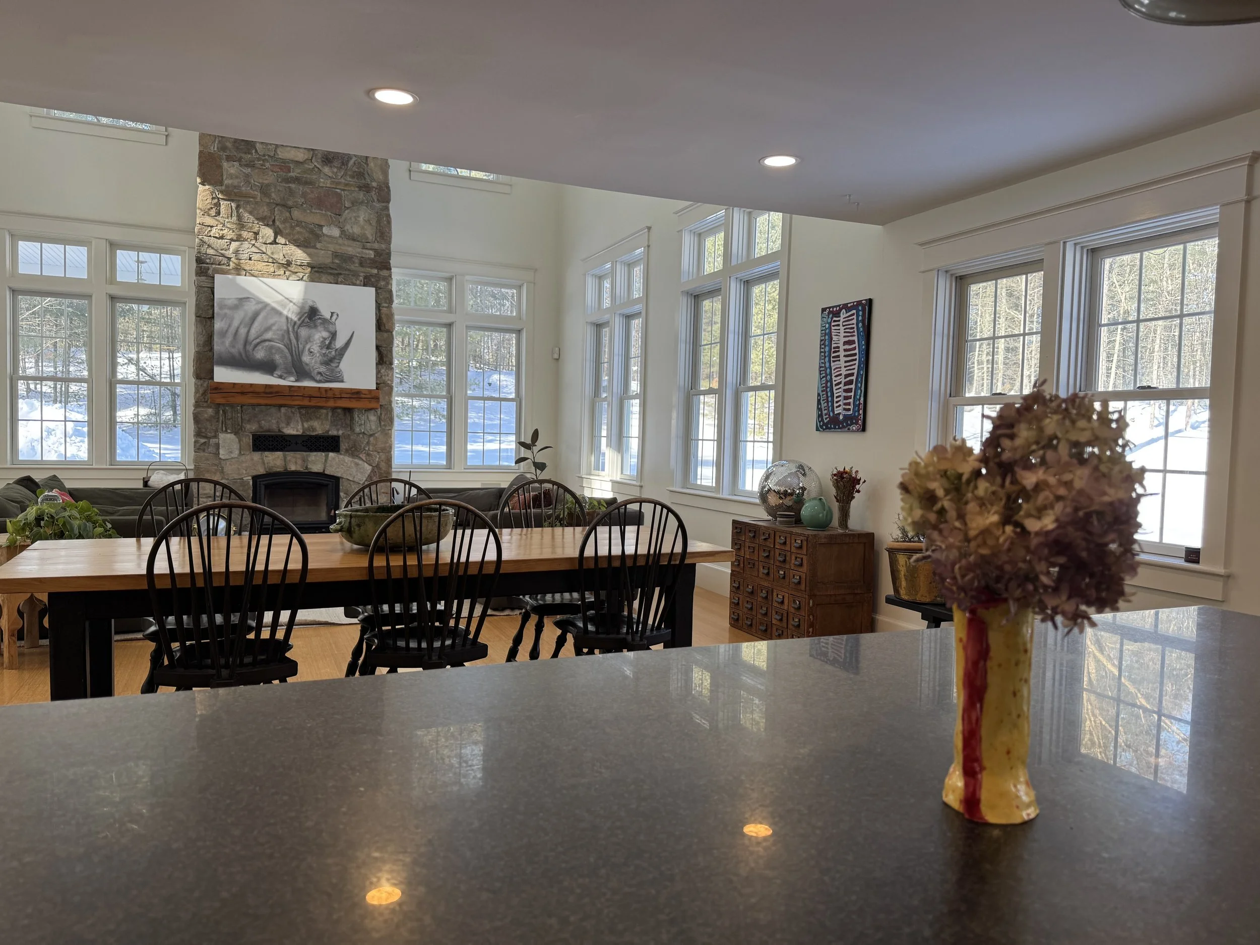 A cozy, well-lit dining area with large windows showing a snowy outdoor landscape, a stone fireplace with a rhino painting above, a long wooden dining table with black chairs, and a colorful flower arrangement on a dark countertop in the foreground.