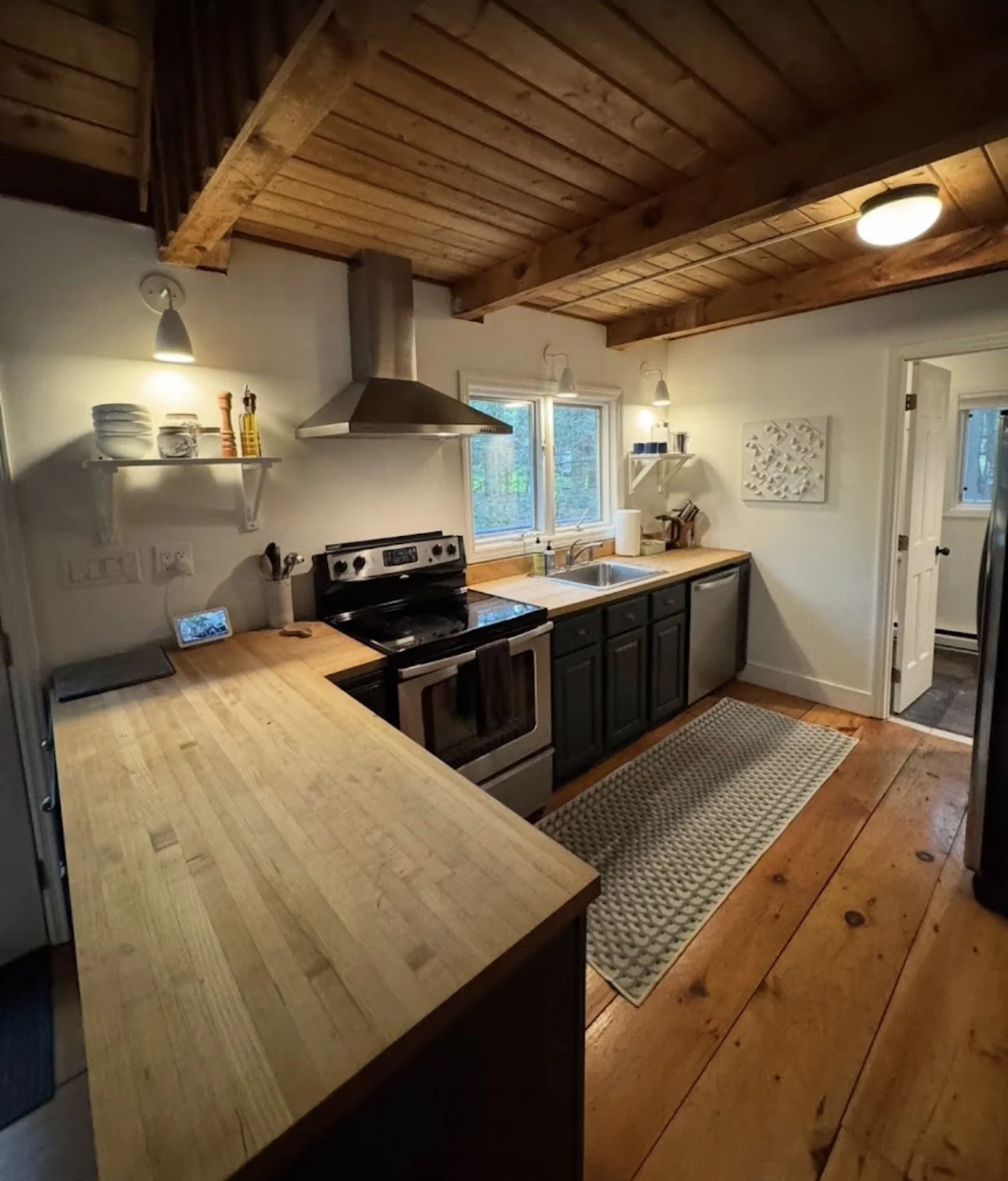 A cozy kitchen with a wooden ceiling and flooring. It has black cabinets, a stainless steel stove with a range hood, and a double window above the sink. There are open shelves with dishes and a few kitchen items. A long wooden countertop extends on t
