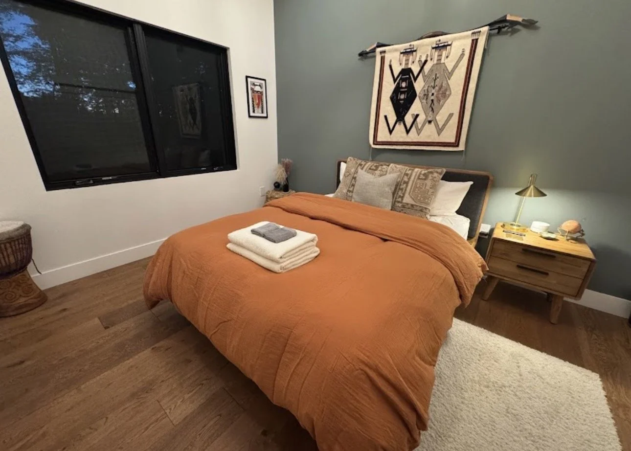 A cozy bedroom with a bed covered in an orange quilt, accompanied by pillows, and a folded towel with toiletries on top. There is a wooden nightstand with a lamp, a small decorative item, and some cups. A window is on the left, with a framed picture 