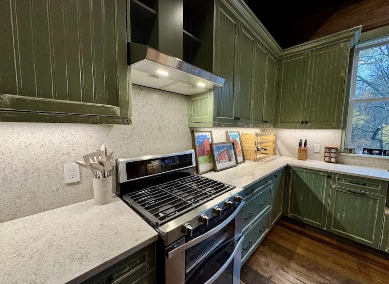 A kitchen with green wooden cabinets, a gas stove, and a window. There are framed pictures, a wooden knife holder, and small jars on the countertops.