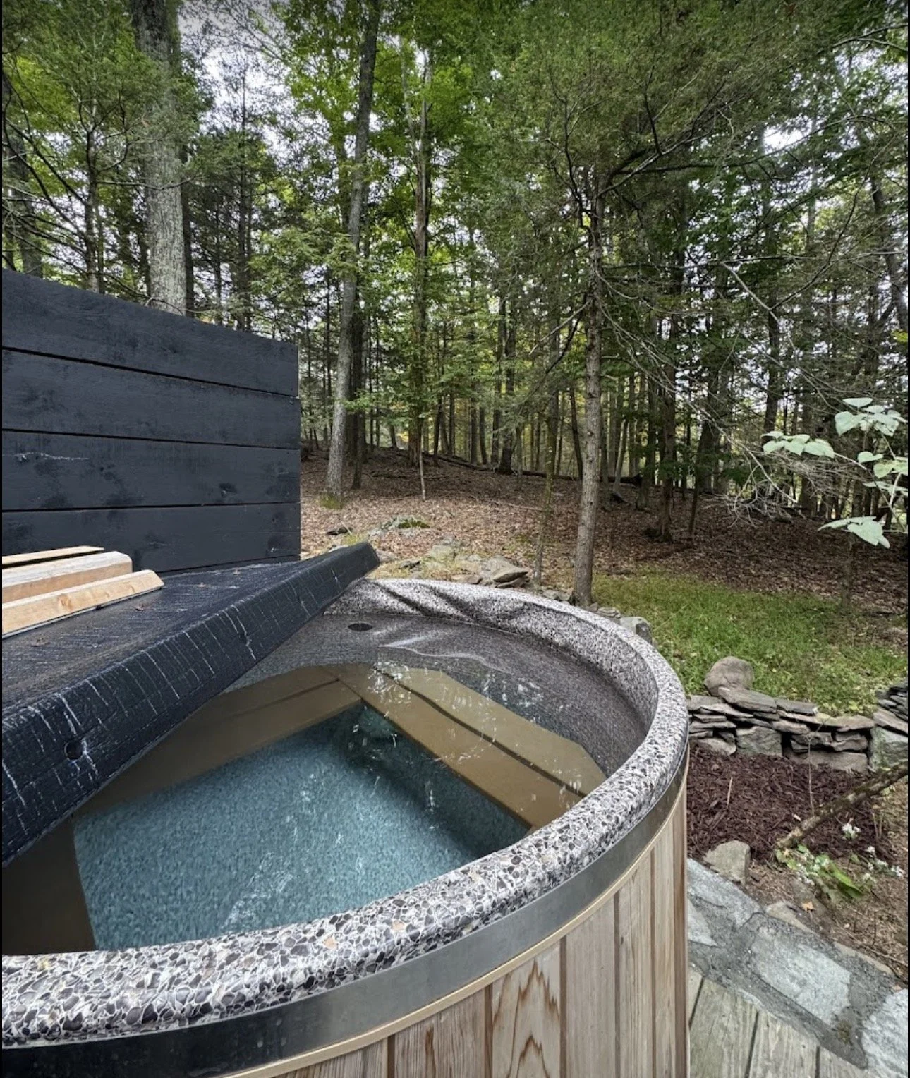 A hot tub with water on a wooden deck in a wooded backyard with trees and rocks.