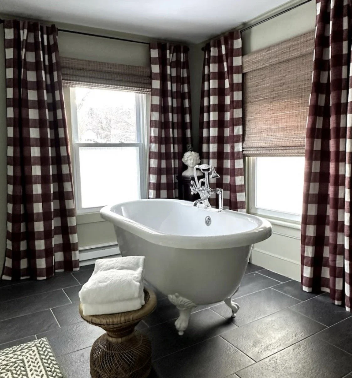 A bathroom with a clawfoot bathtub positioned in front of two windows with checkered red and white curtains and woven blinds, a small stool with rolled white towels, and a decorative bust sculpture on a pedestal.