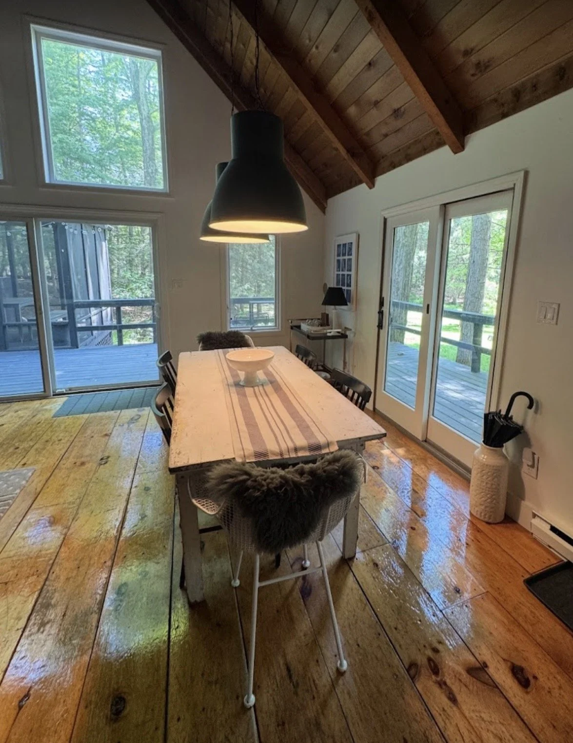 Dining room with wooden floors, a large wooden table with chairs, sliding glass door leading to a deck, large windows showing trees outside, and a sloped wooden ceiling.