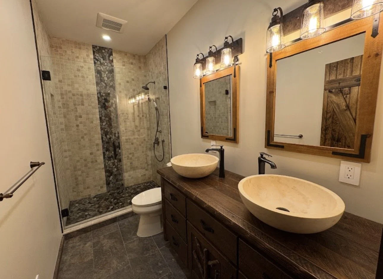 Bathroom with a double vanity featuring two vessel sinks and two mirrors with wooden frames, a wall-mounted light fixture, a walk-in shower with beige and pebble tile, and a toilet.