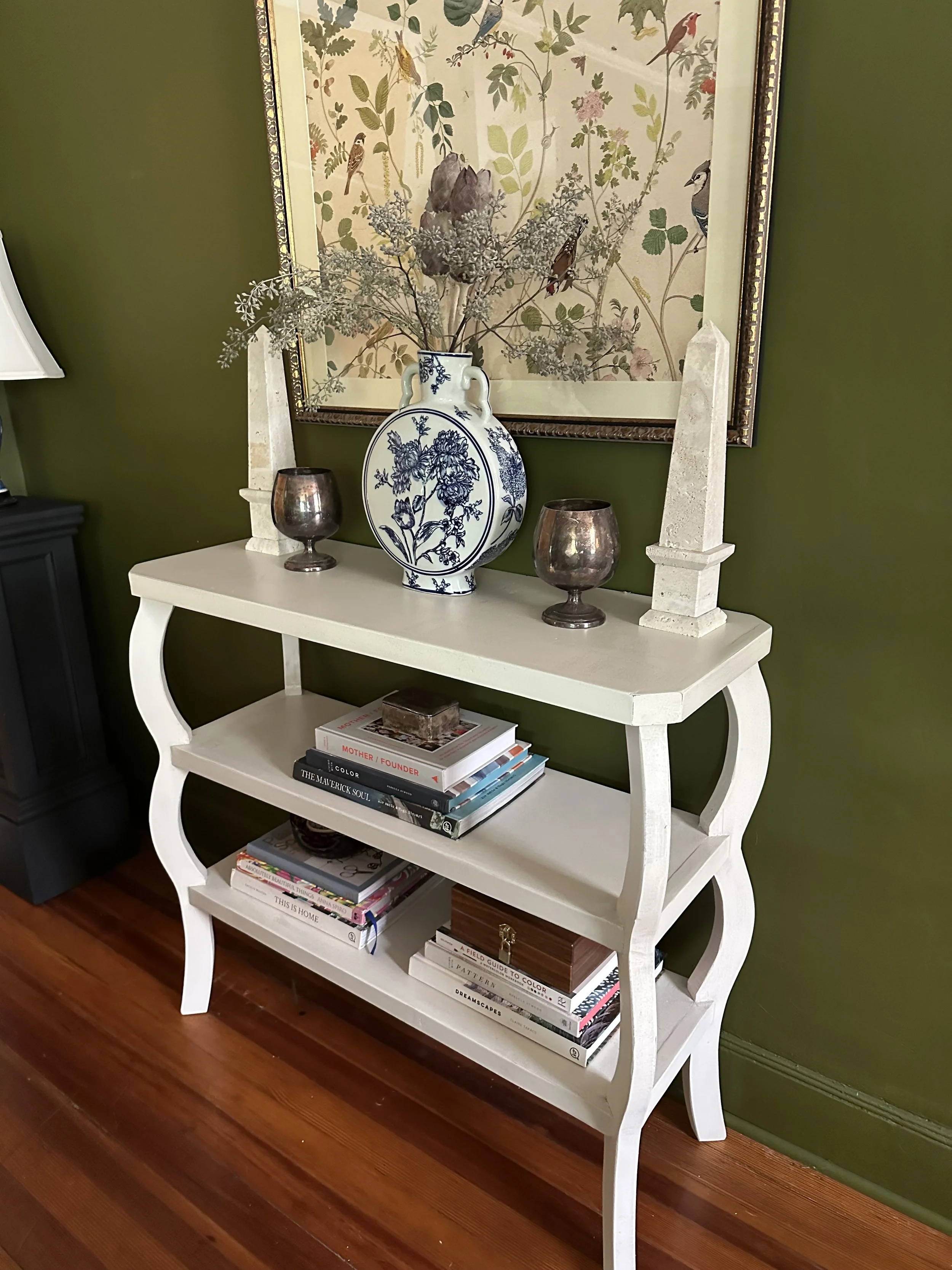 A white wooden bookshelf with three shelves, decorated with a large blue and white ceramic vase filled with dried flowers, two metallic candle holders, and two white obelisk decorations on top. A framed botanical illustration hangs on a green wall be
