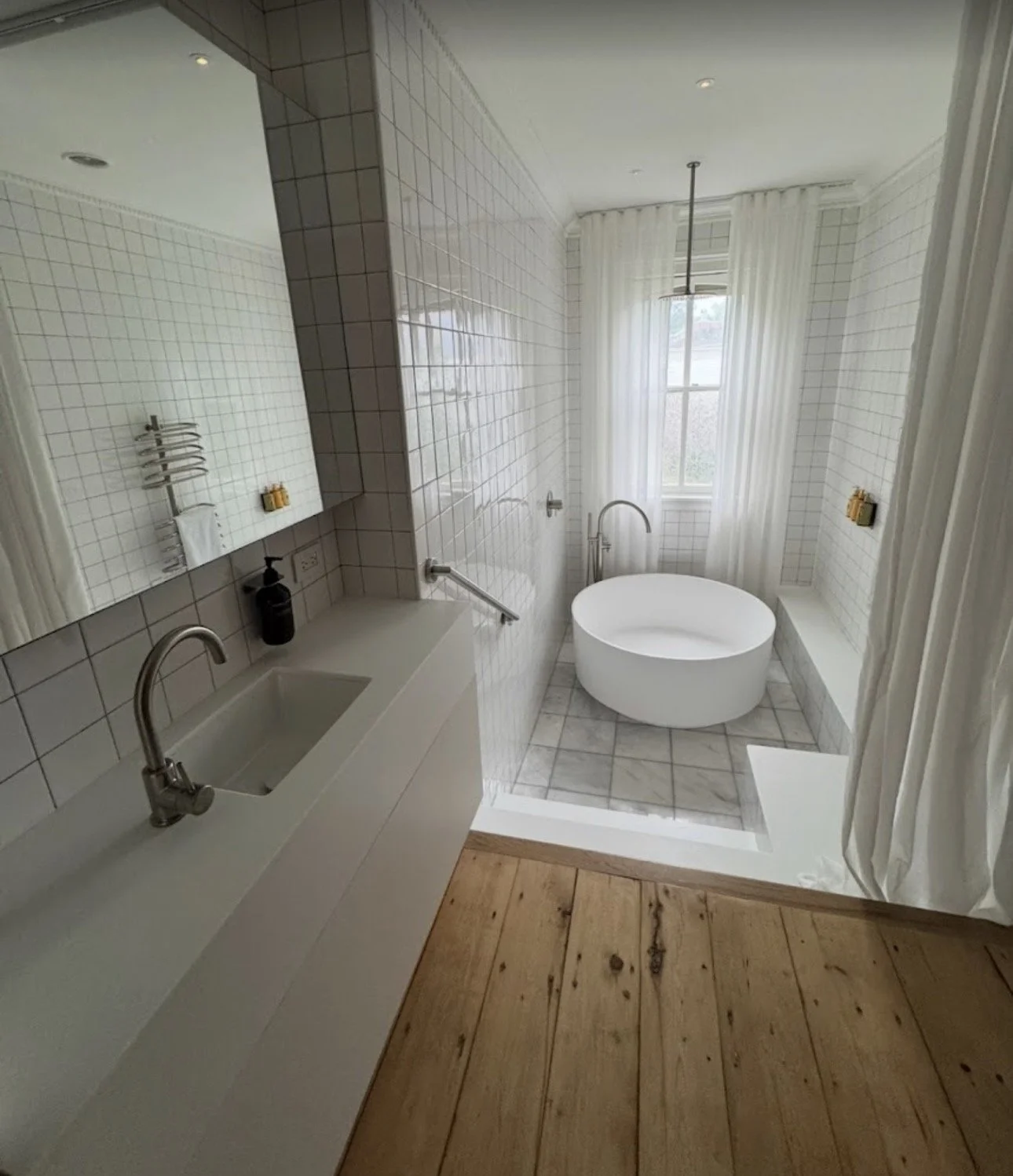 A modern bathroom with white tiled walls and a wooden floor. It features a small sink with a black soap dispenser, a large mirror, and a circular bathtub near a window with white curtains. There is a shower with a ceiling-mounted showerhead and a sma