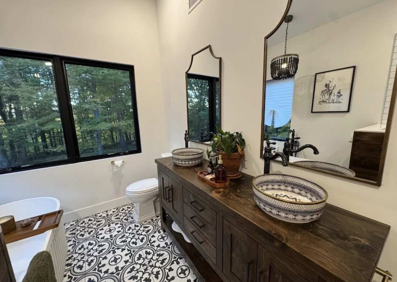 Bathroom with large window showing trees outside, dark wooden vanity with two patterned vessel sinks, large mirrors, potted plant, decorative items, black vintage-style faucets, patterned tile floor, wall art, and hanging light fixture.
