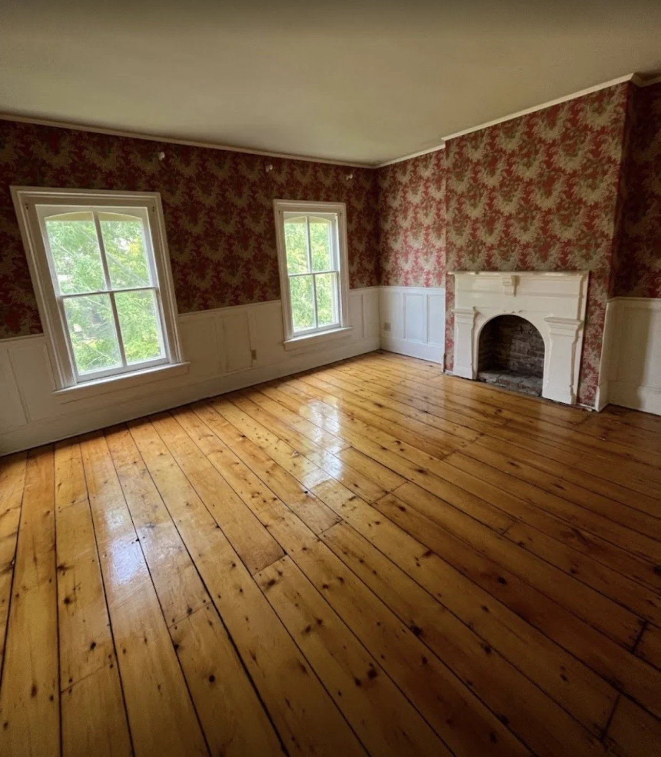 Empty room with two windows, a white fireplace, wood flooring, and floral wallpaper on one wall.