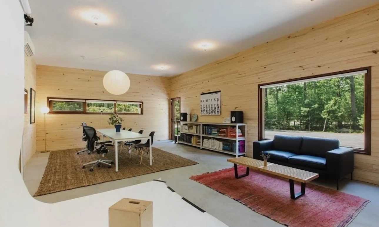 Bright living and dining room with light wood paneled walls, large windows showing greenery outside, a black sofa on a red rug, a white bookshelf with books and speakers, a rectangular coffee table, a white dining table with six black chairs, and a p